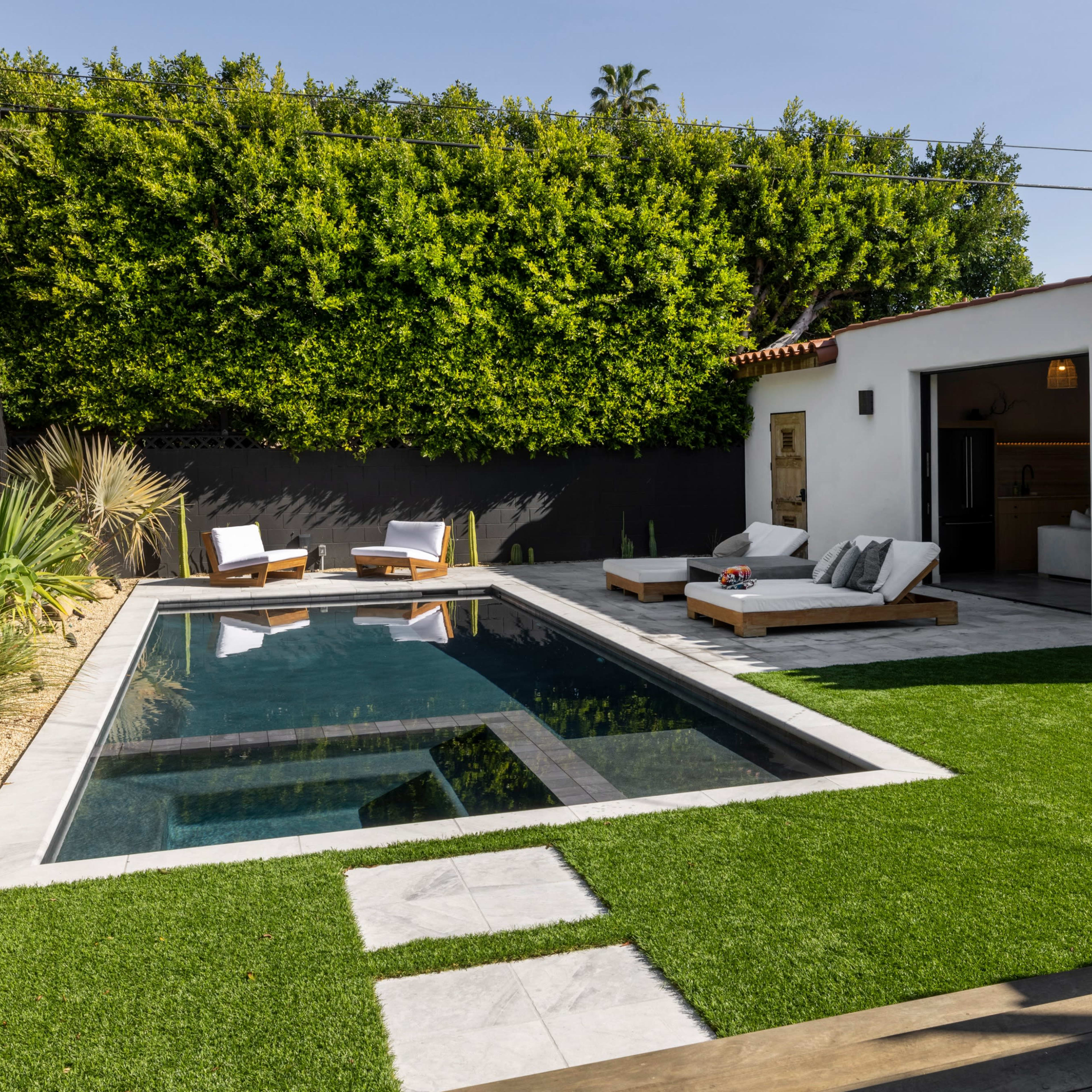 A modern outdoor space featuring a rectangular swimming pool surrounded by lounge chairs, artificial turf, and a wall of greenery.