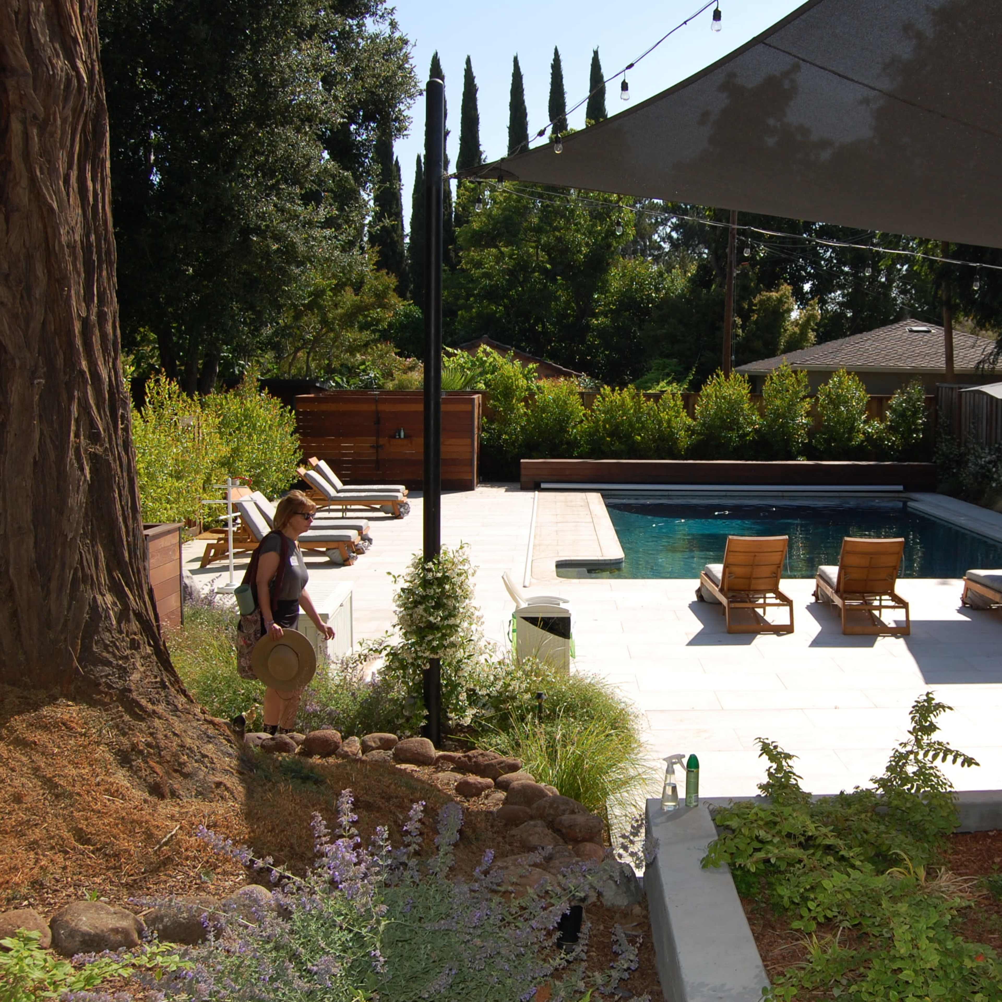 A person carrying a straw hat walks along a pathway beside a modern pool surrounded by lounge chairs and landscaped greenery.