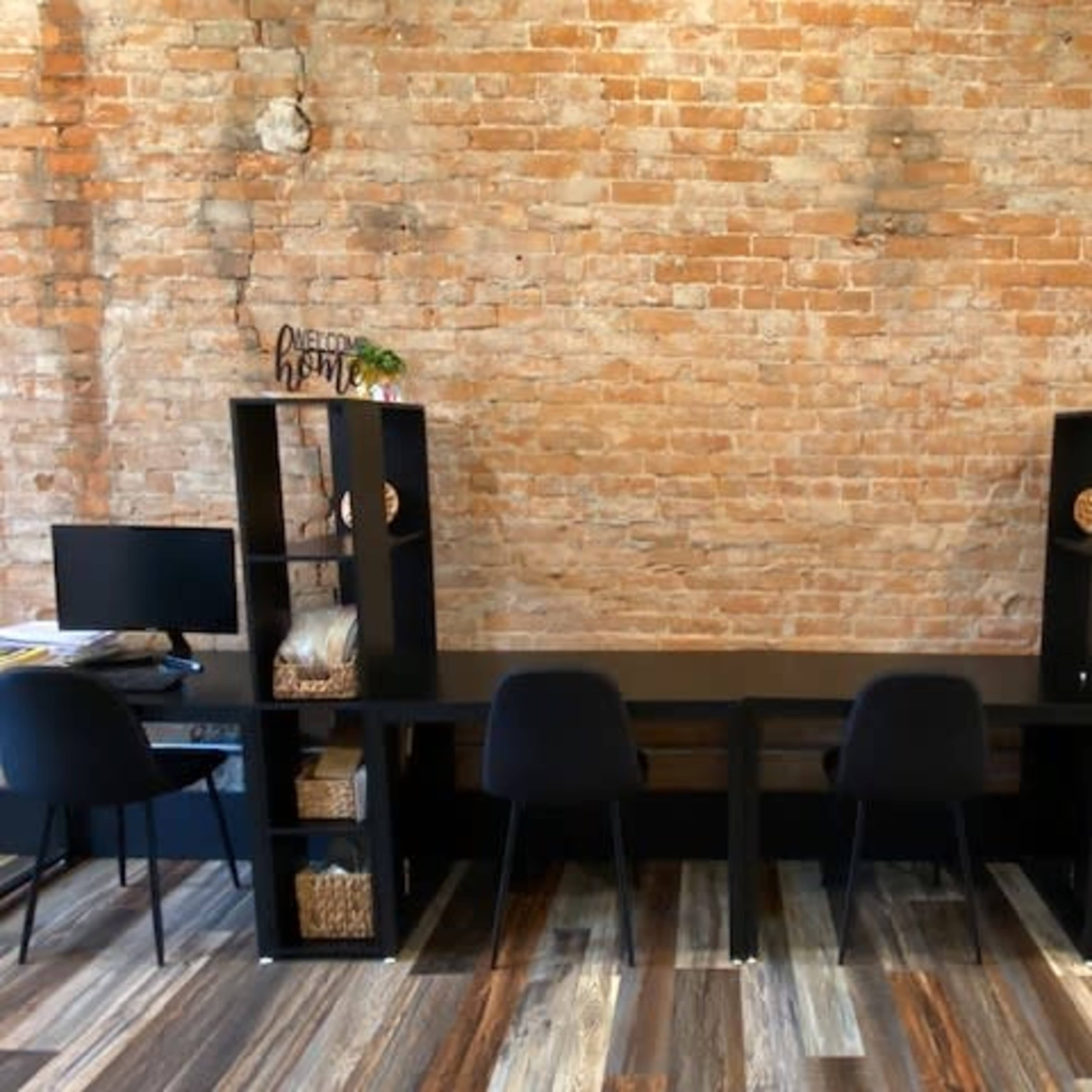 A long, black wooden desk with black chairs is set against a rustic brick wall, flanked by two black shelving units containing baskets and decorative items.