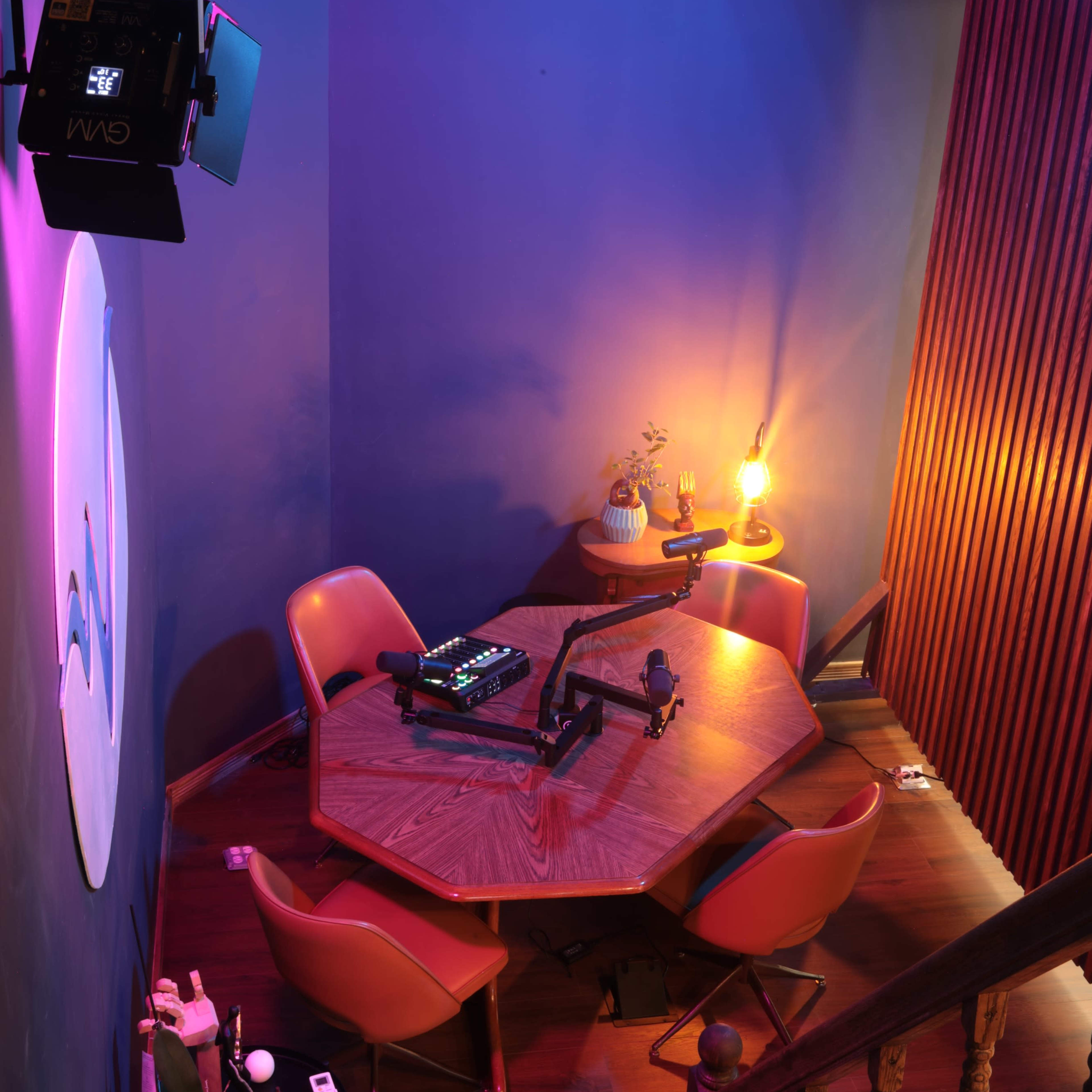 A dimly lit studio features a hexagonal wooden table surrounded by four orange chairs, with a spotlight and decorative items on display.