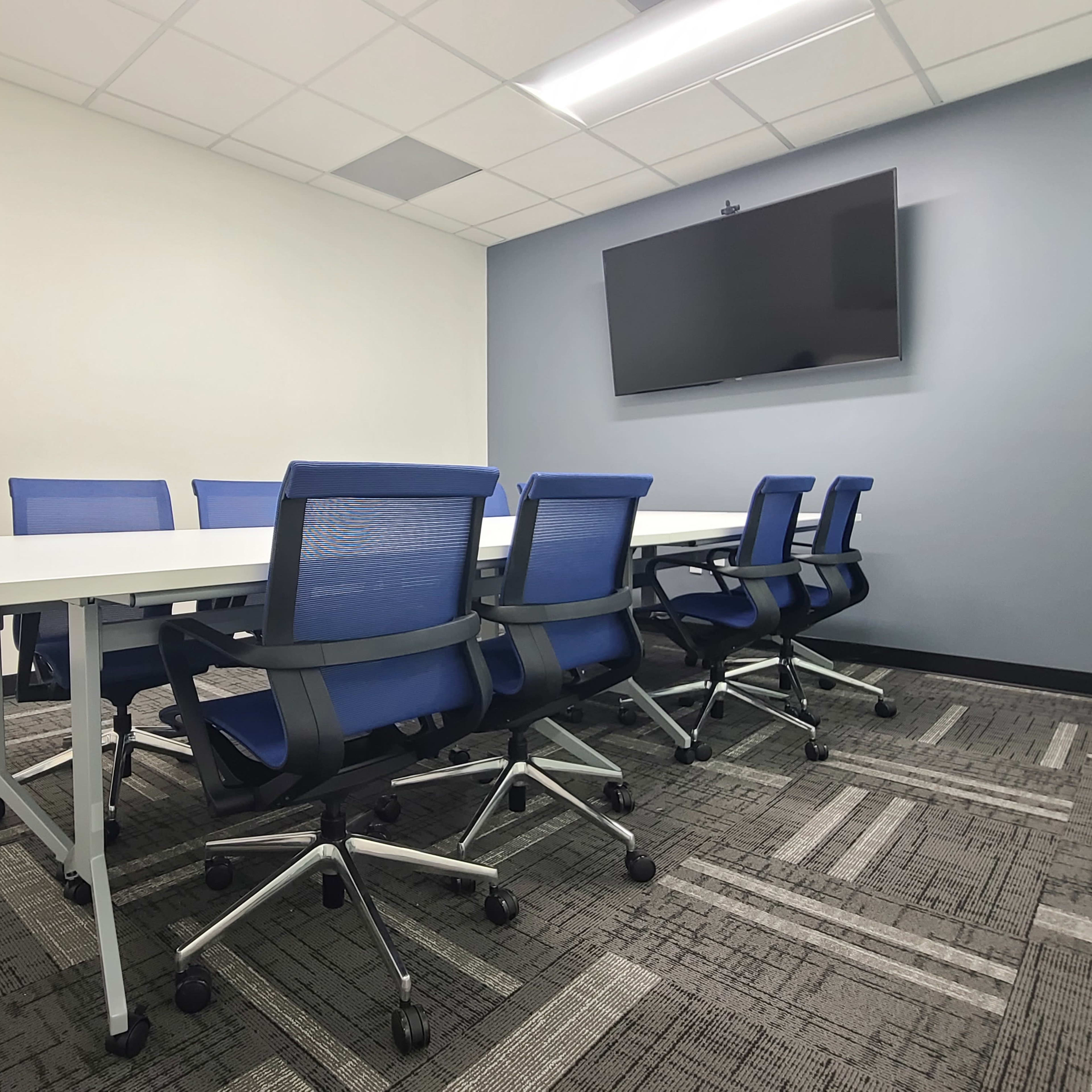 A modern conference room features a long white table surrounded by blue ergonomic chairs and a wall-mounted television.