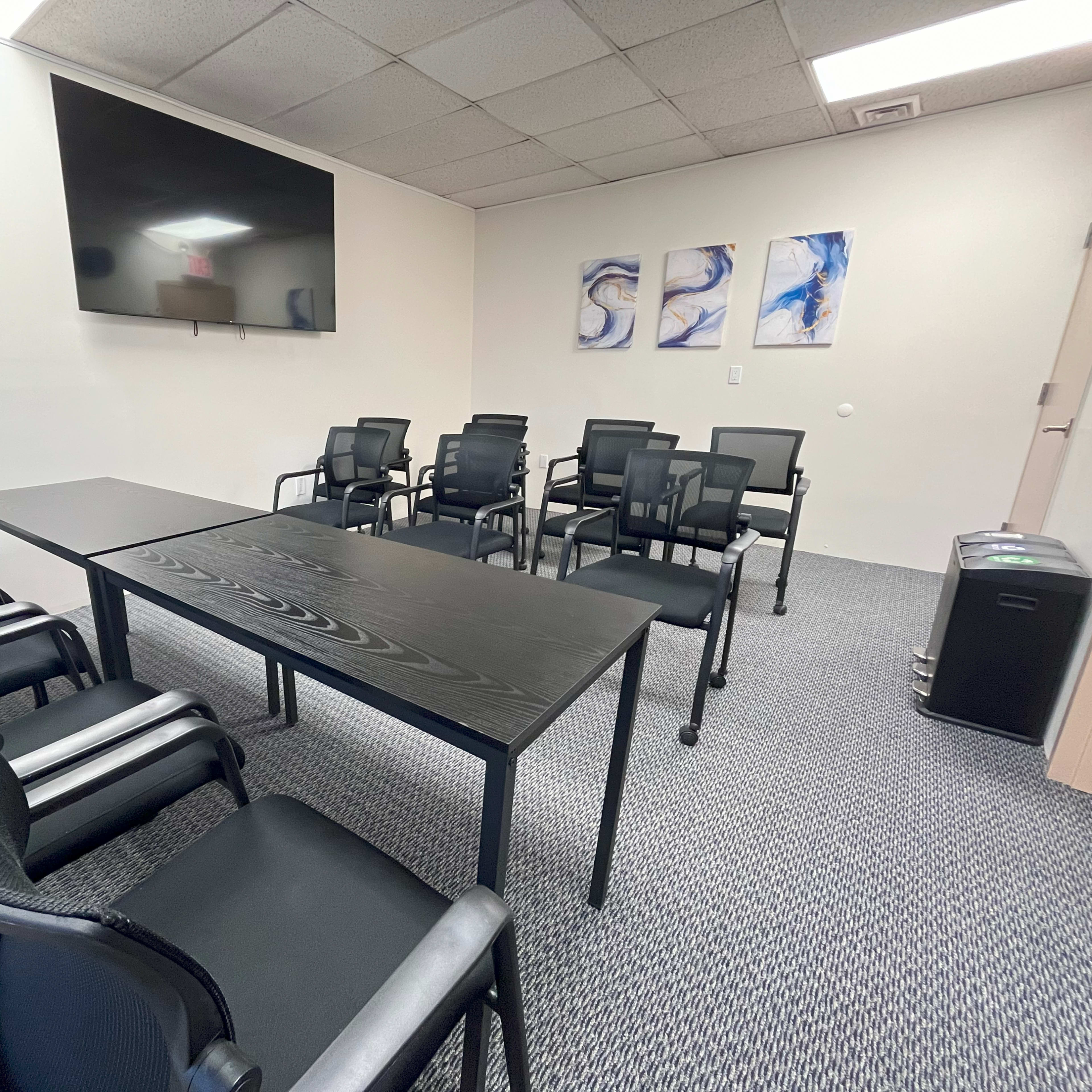 A conference room features a long black table surrounded by multiple black chairs, with a large screen mounted on the wall and abstract artwork displayed nearby.