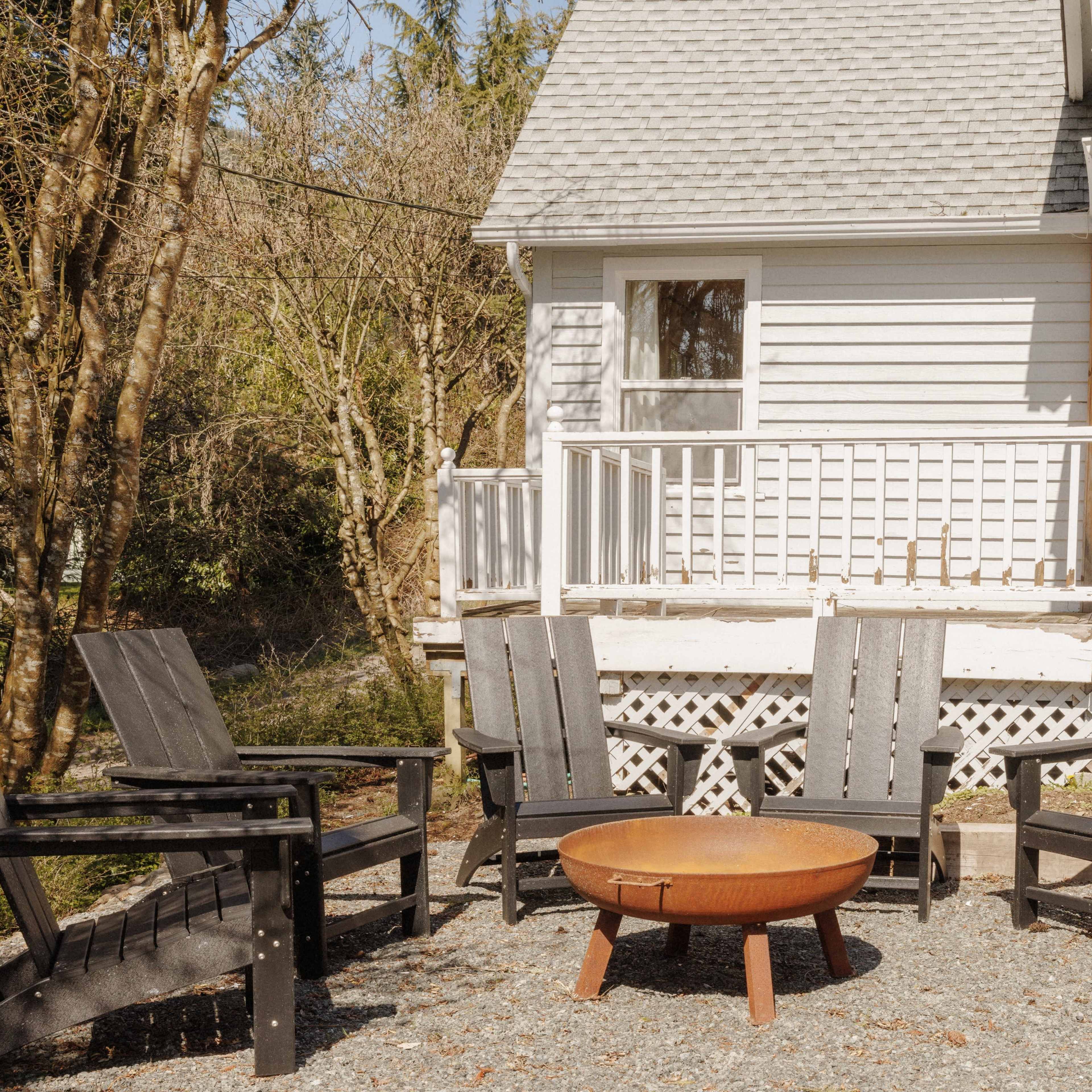 A set of four black Adirondack chairs surrounds a circular fire pit on a gravel area next to a house with a white exterior and a porch.