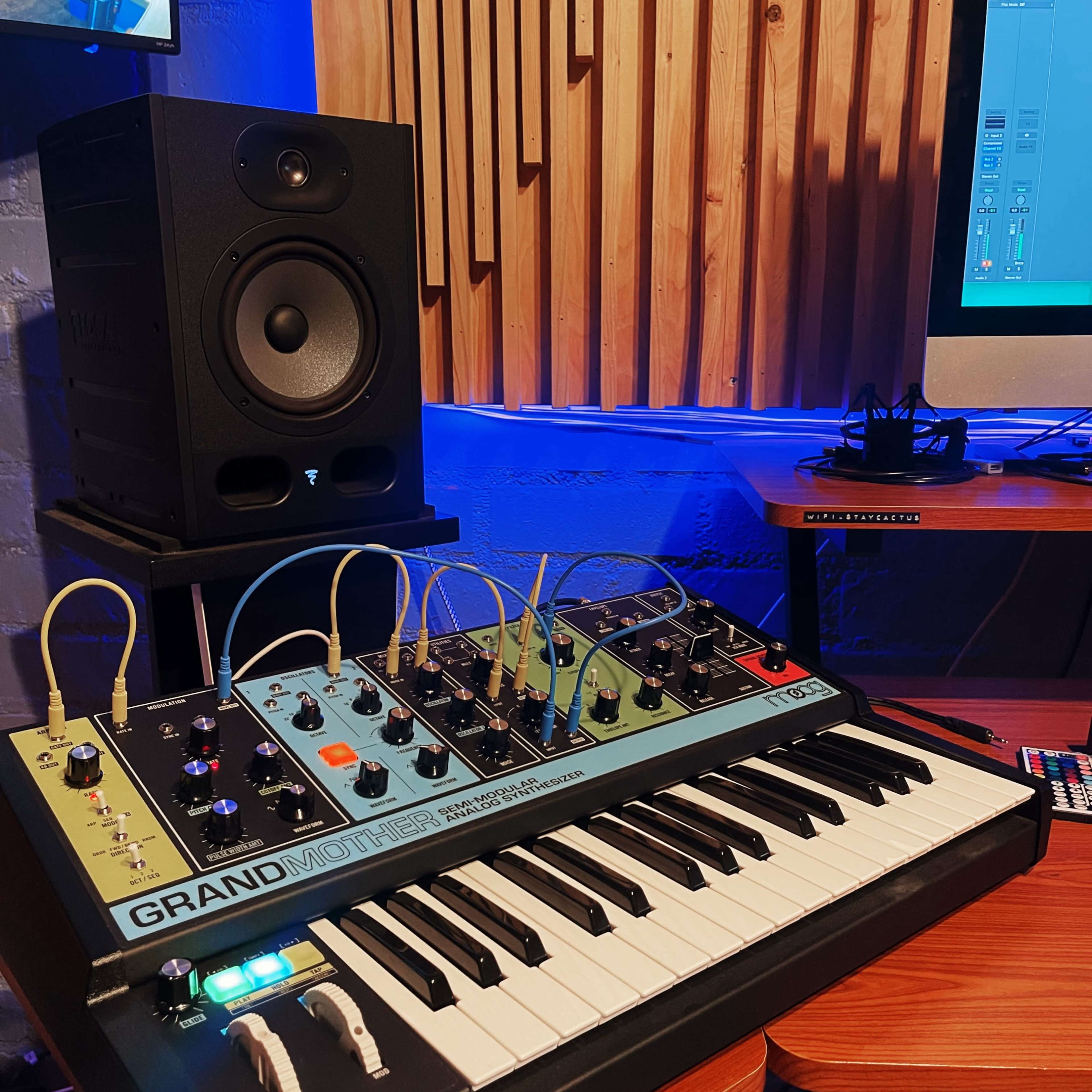 A synthesizer with a keyboard and various knobs is situated in front of a studio monitor and a computer screen displaying audio software, surrounded by wooden acoustic panels.