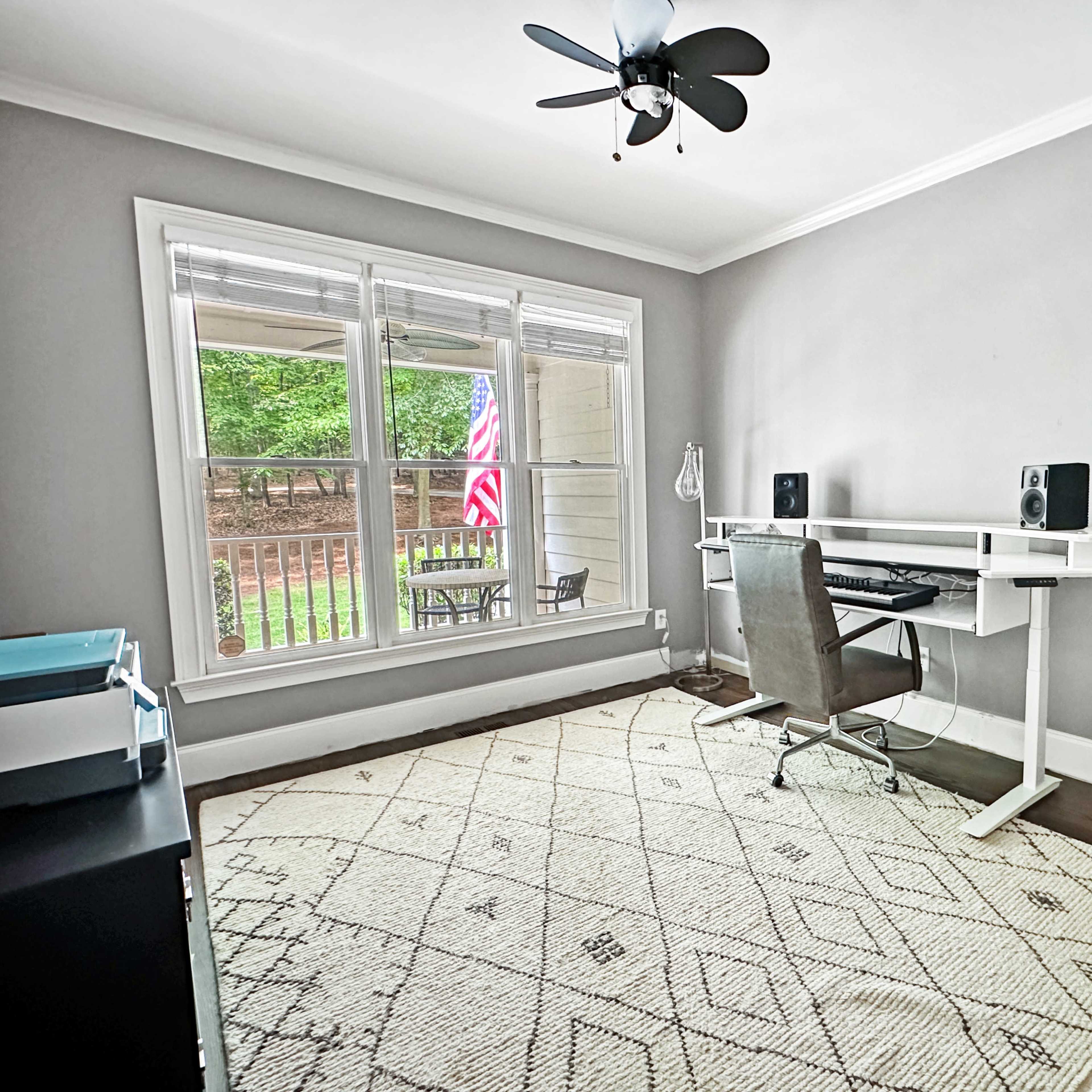 A minimalistic office space features a desk with a chair, speakers, and a window overlooking a patio with an American flag visible outside.