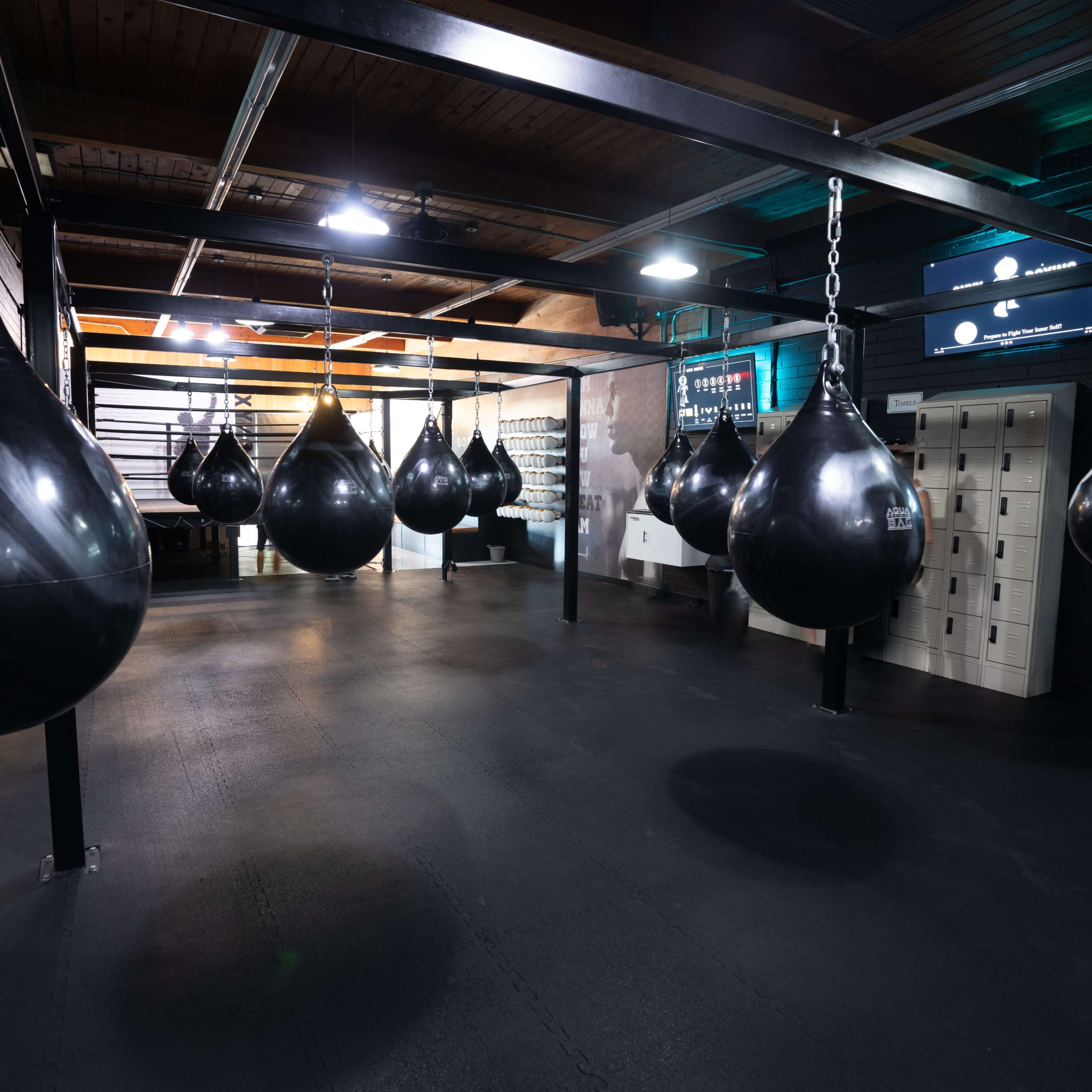 A gym interior featuring several black punching bags hanging from a metallic frame in a spacious area with dark flooring.