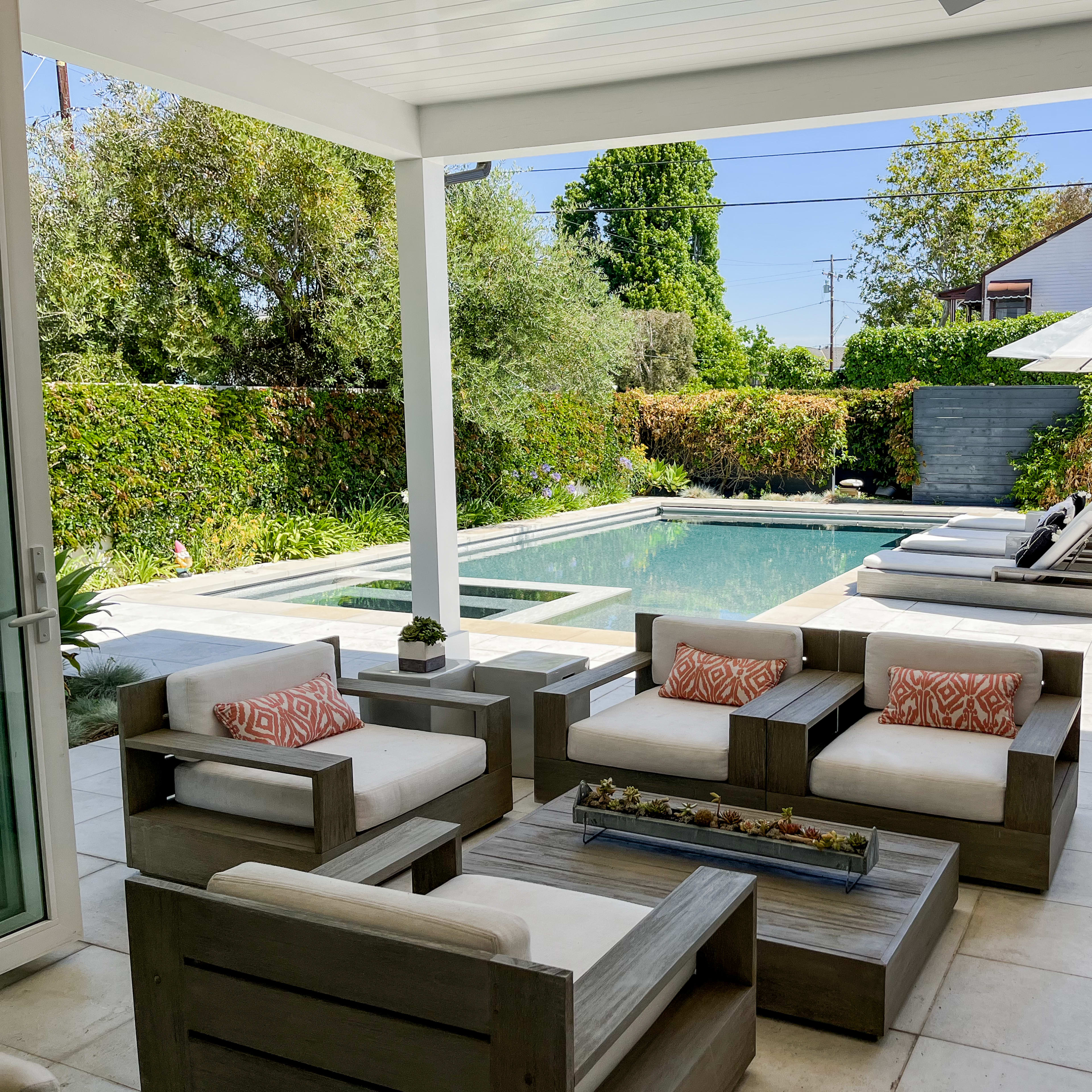 A modern outdoor patio features comfortable seating, a rectangular swimming pool, and a shaded umbrella area surrounded by lush greenery.