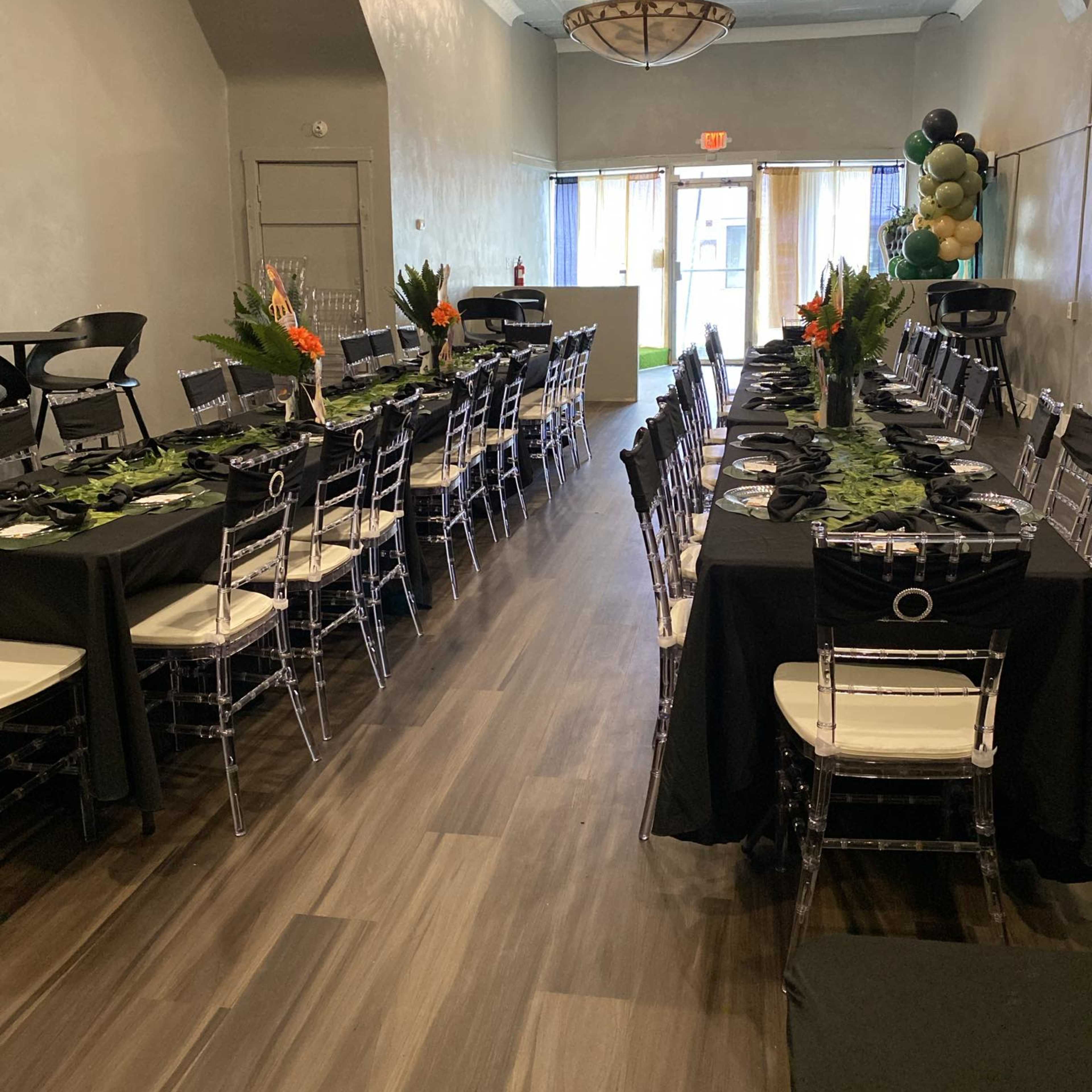 A decorated dining area features long tables set with black tablecloths, clear chairs, and tropical-themed centerpieces, arranged symmetrically along the space.