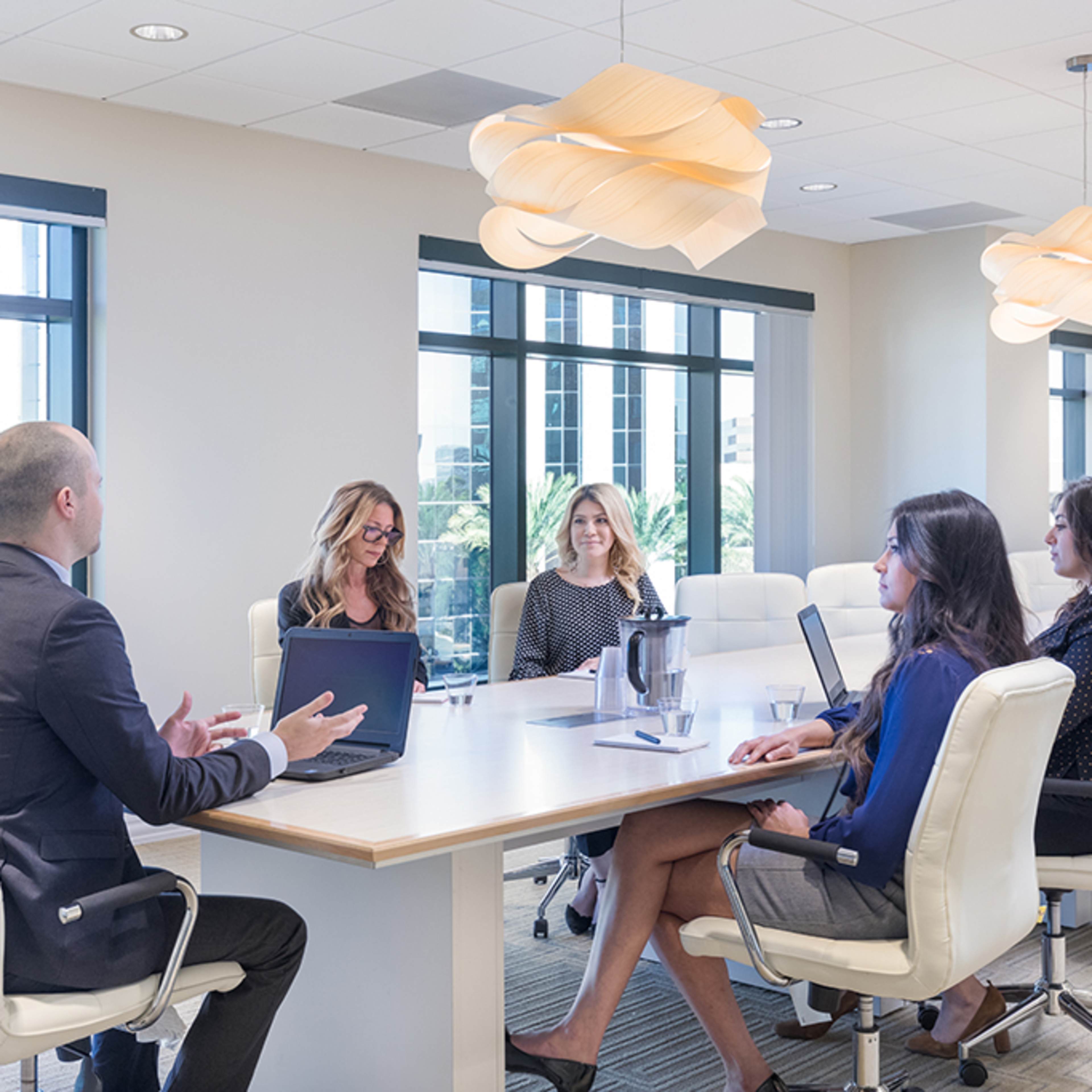 A group of five professionals engages in a discussion around a modern conference table in a well-lit office with large windows.