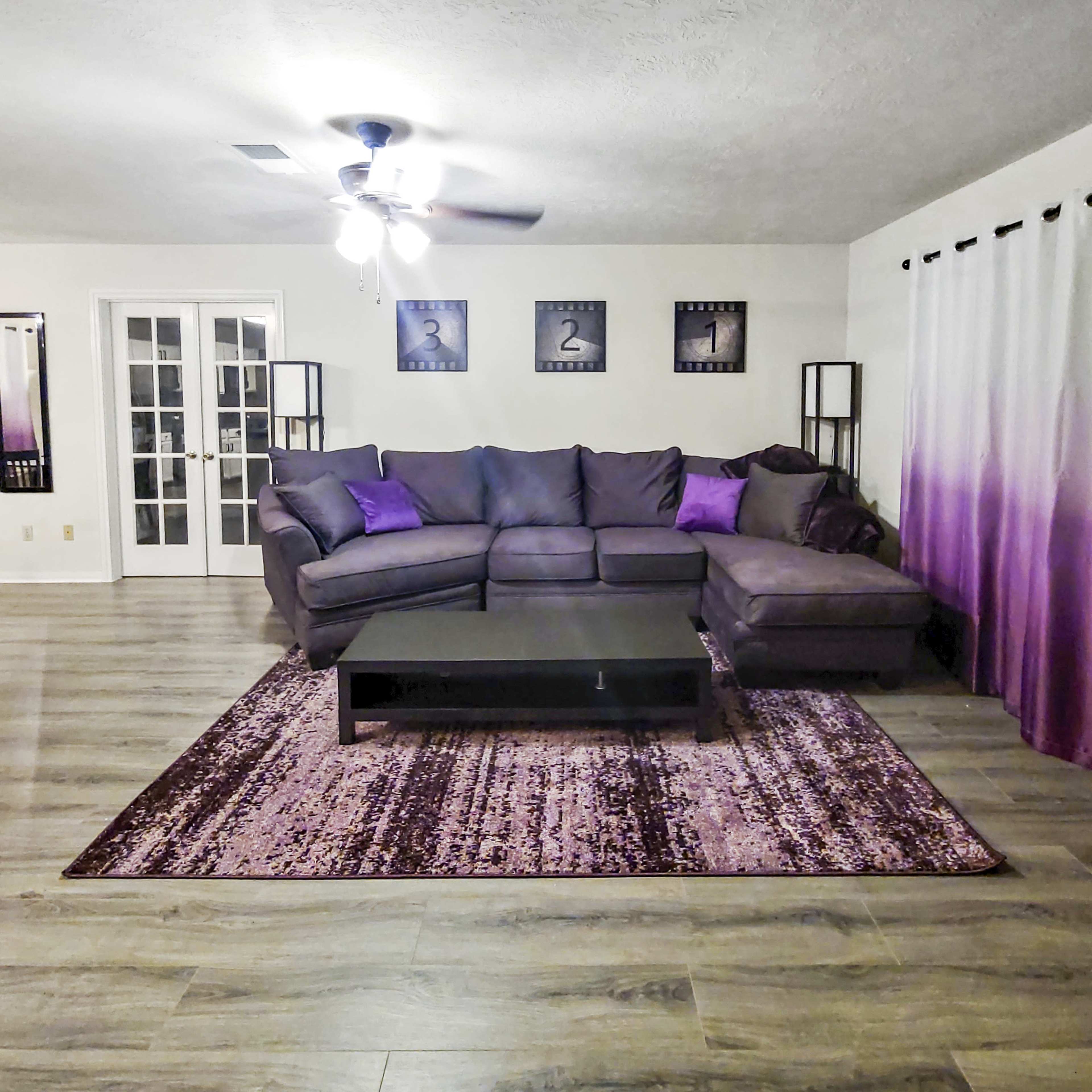 A living room features a purple sectional sofa, a black coffee table, and a patterned rug, with wall art displaying numbers and curtains blending from white to purple.
