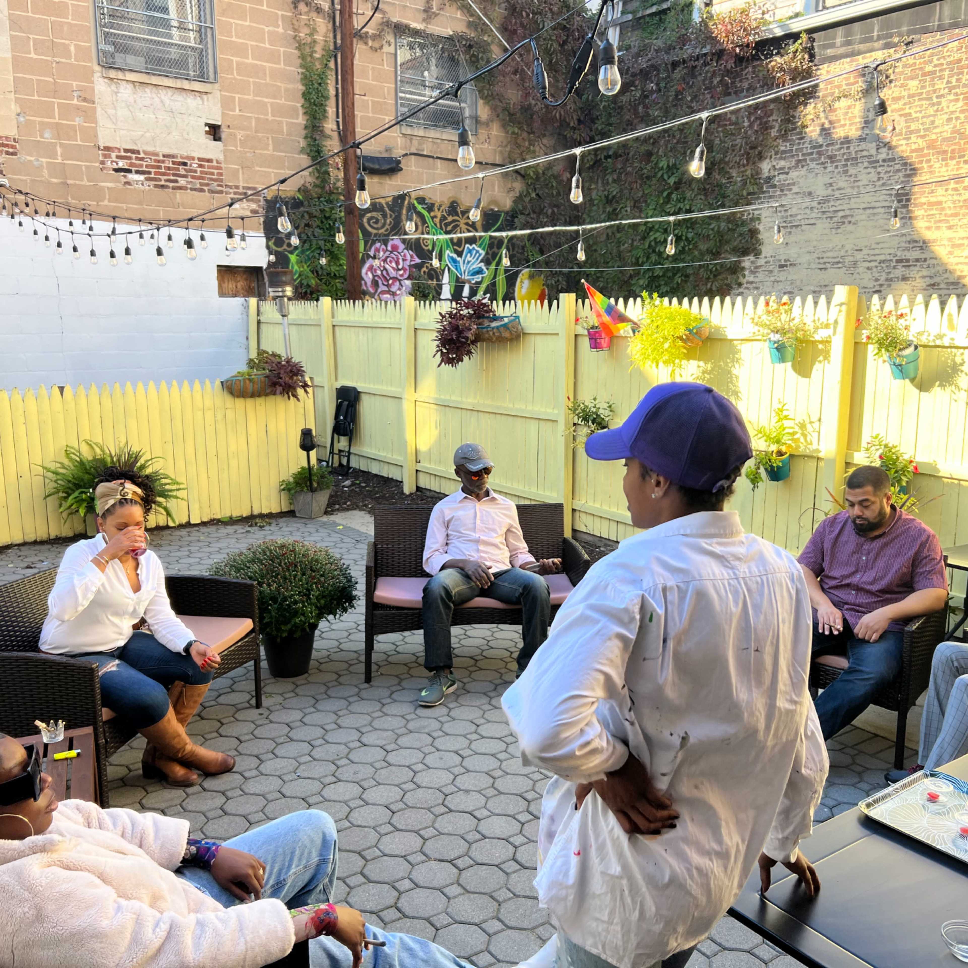 A group of people sits around a patio with string lights and potted plants.