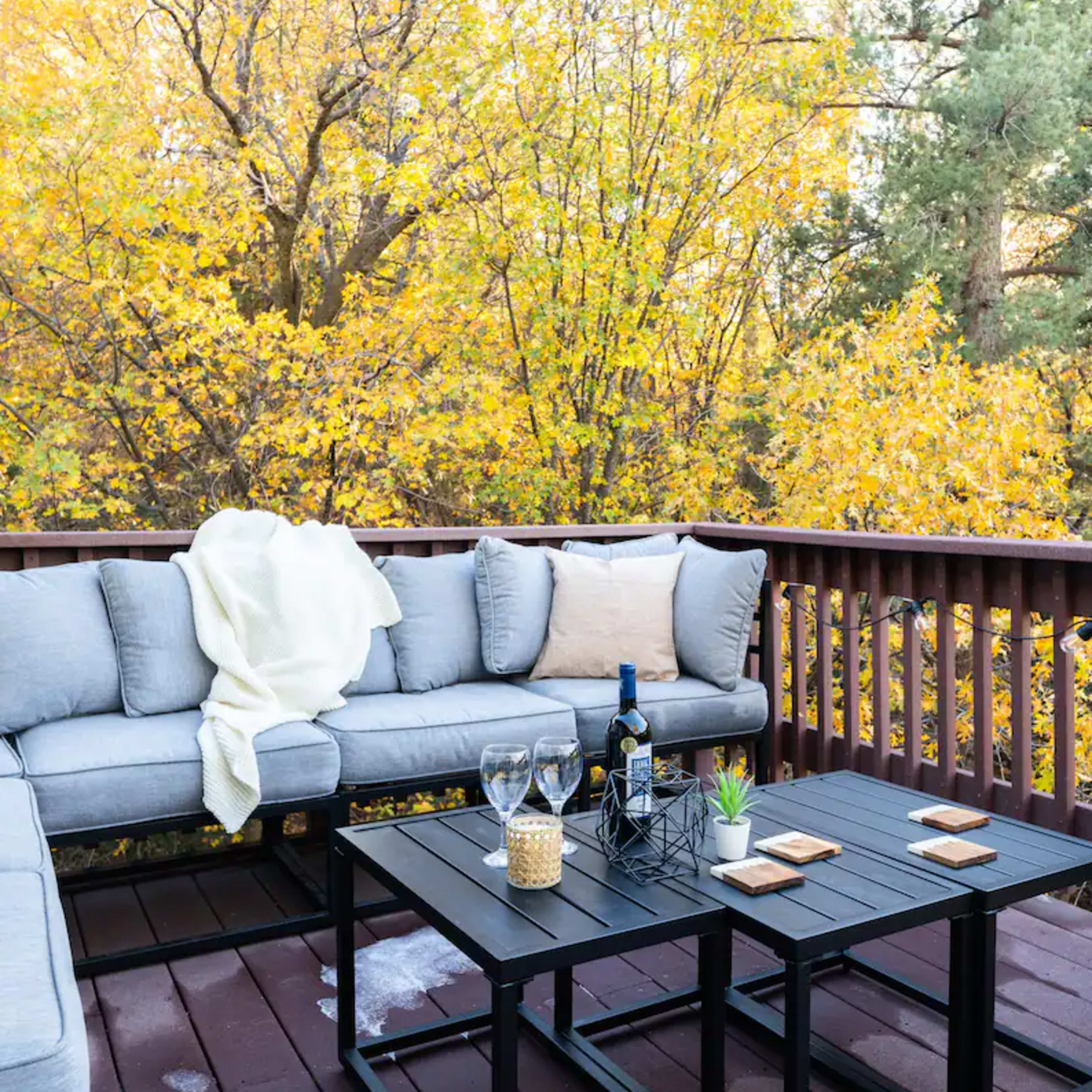 A cozy outdoor seating area features a gray sectional sofa with cushions and a black table surrounded by vibrant autumn foliage.