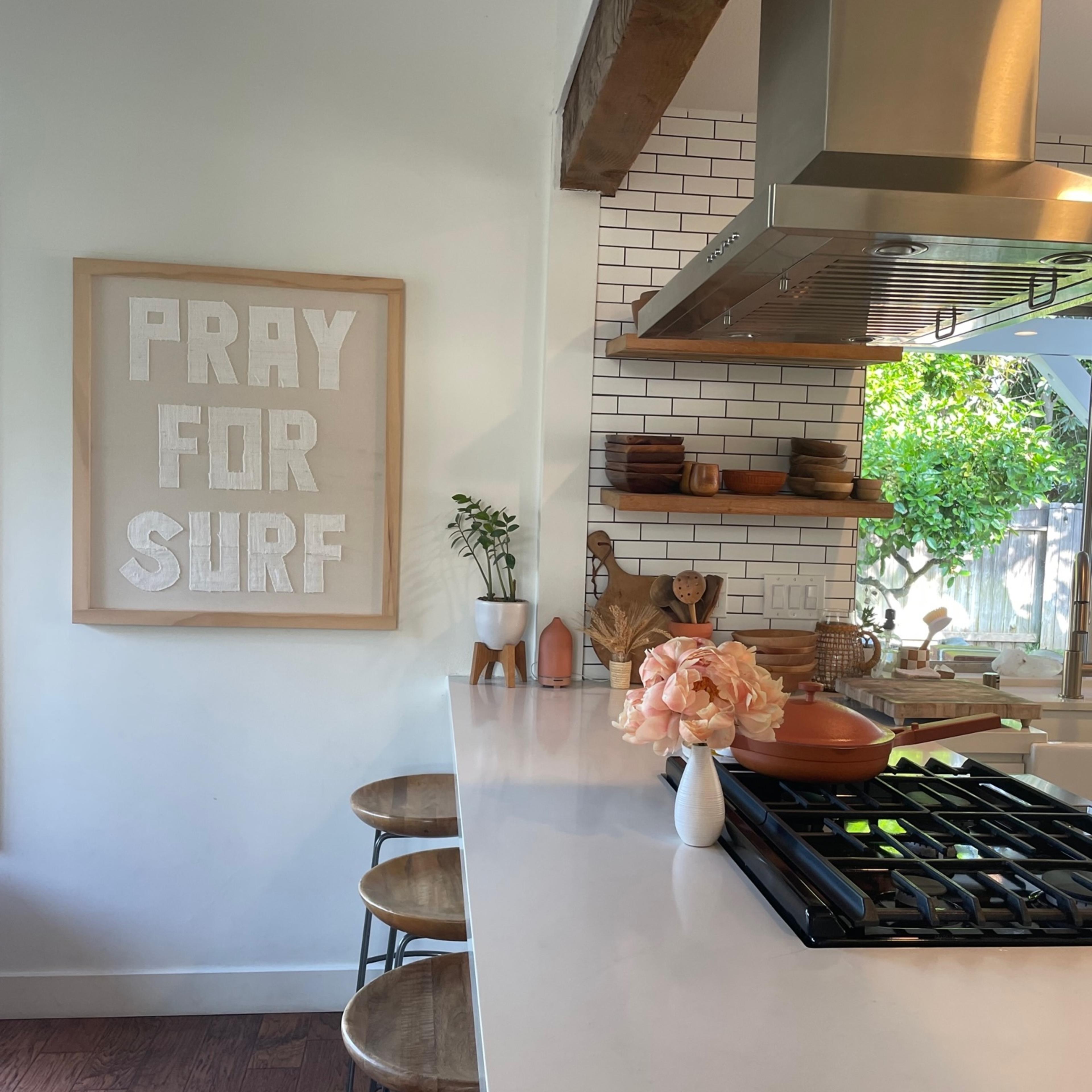 A modern kitchen features a white countertop with bar stools, a wall art piece that reads "PRAY FOR SURF," and a view of greenery through a window.