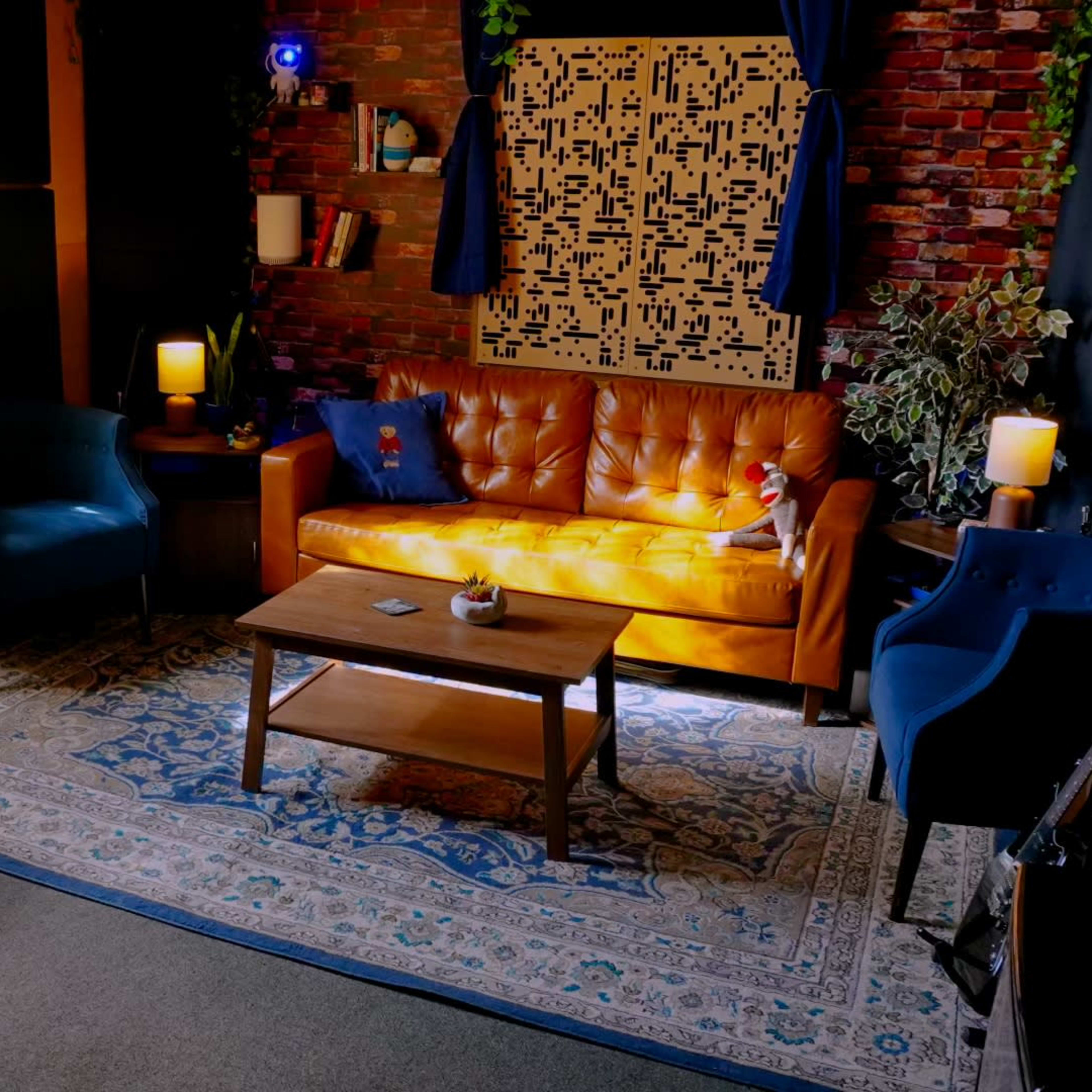 A cozy living room with a brick wall, orange sofa, blue chairs, a coffee table, and a patterned rug under soft lighting.