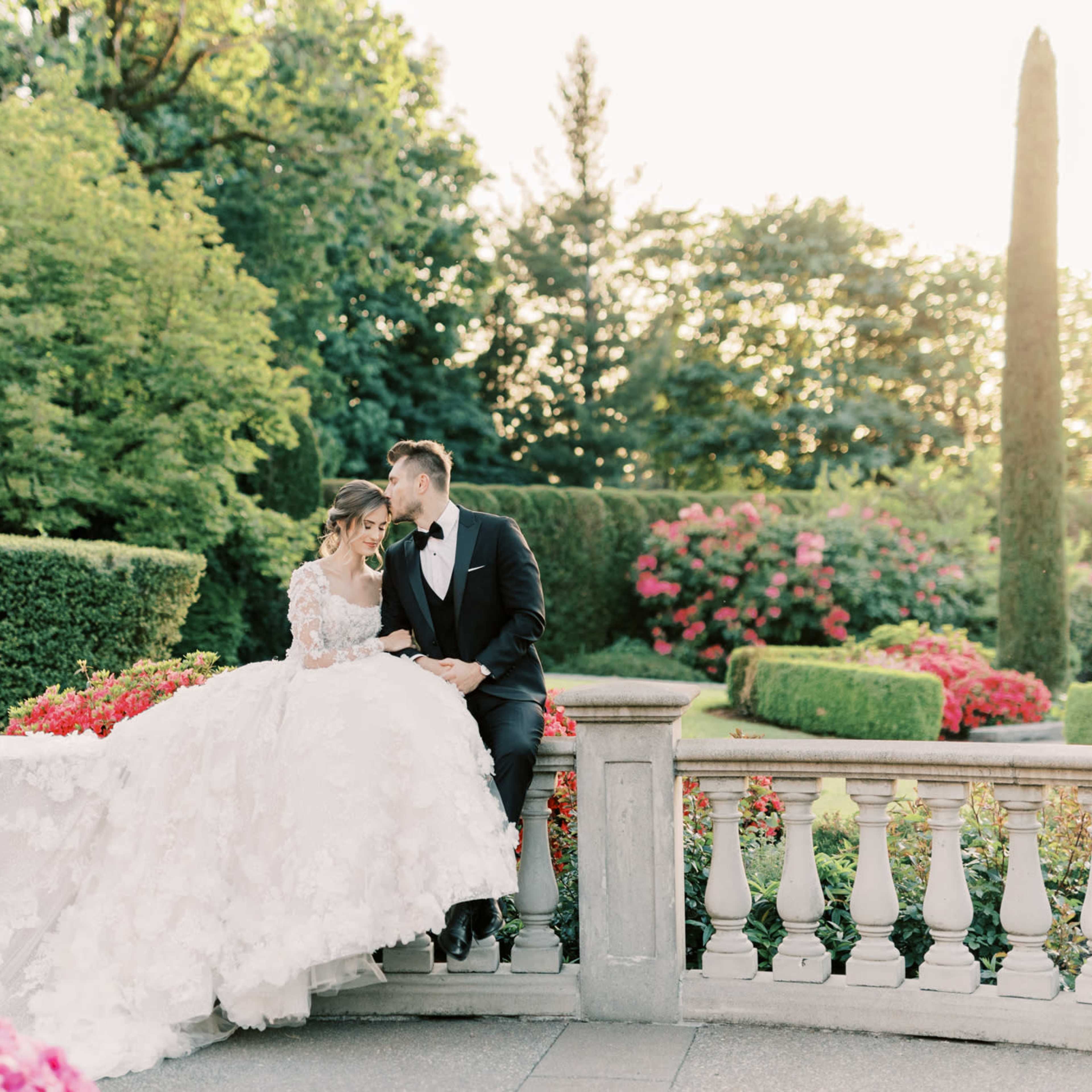 A couple in formal attire sits on a stone railing surrounded by lush gardens and vibrant floral arrangements.