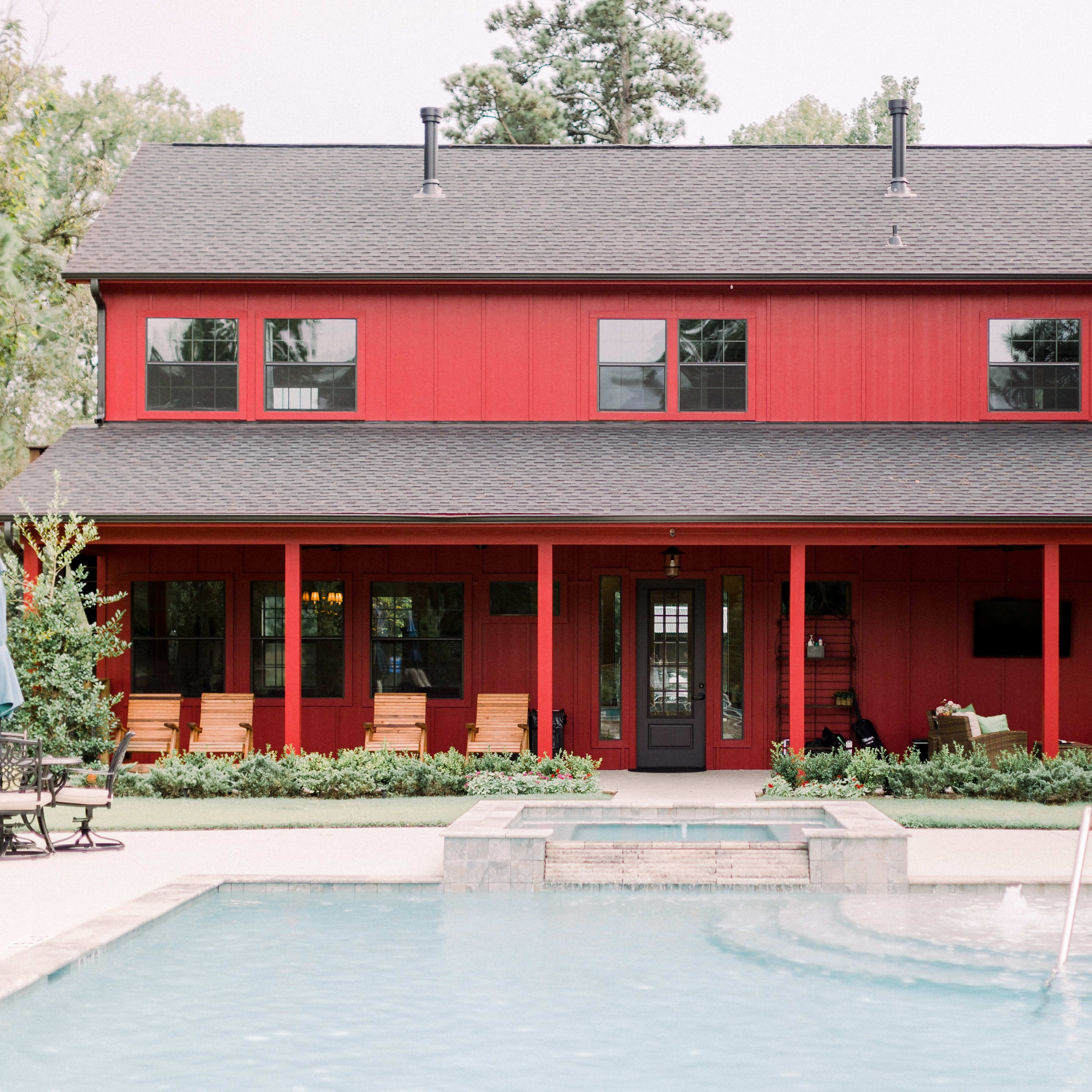 A two-story red house with a covered porch is situated next to a swimming pool surrounded by patio furniture and umbrellas.