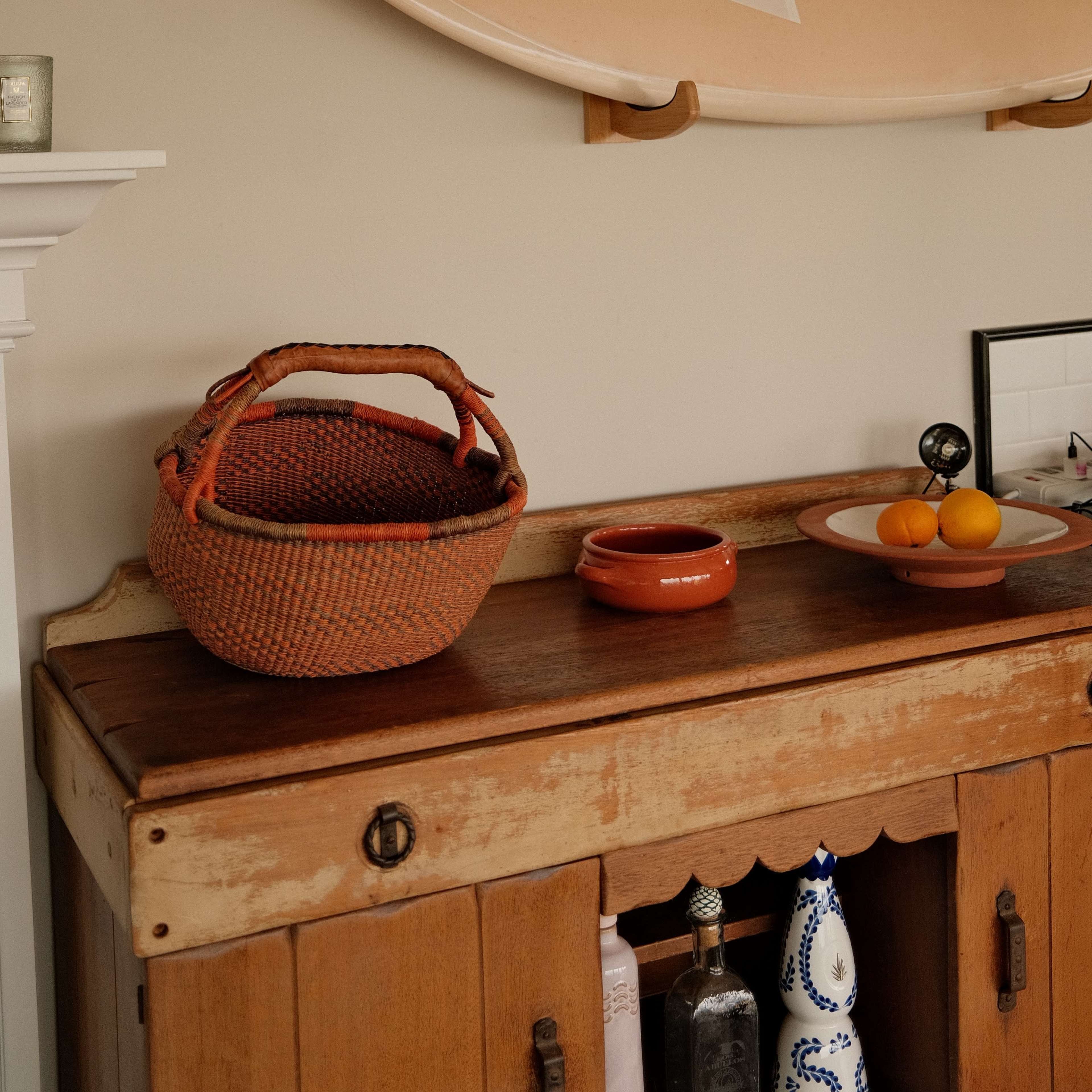 A wooden sideboard displays a woven basket, a small bowl, and fresh oranges beneath a surfboard mounted on the wall.