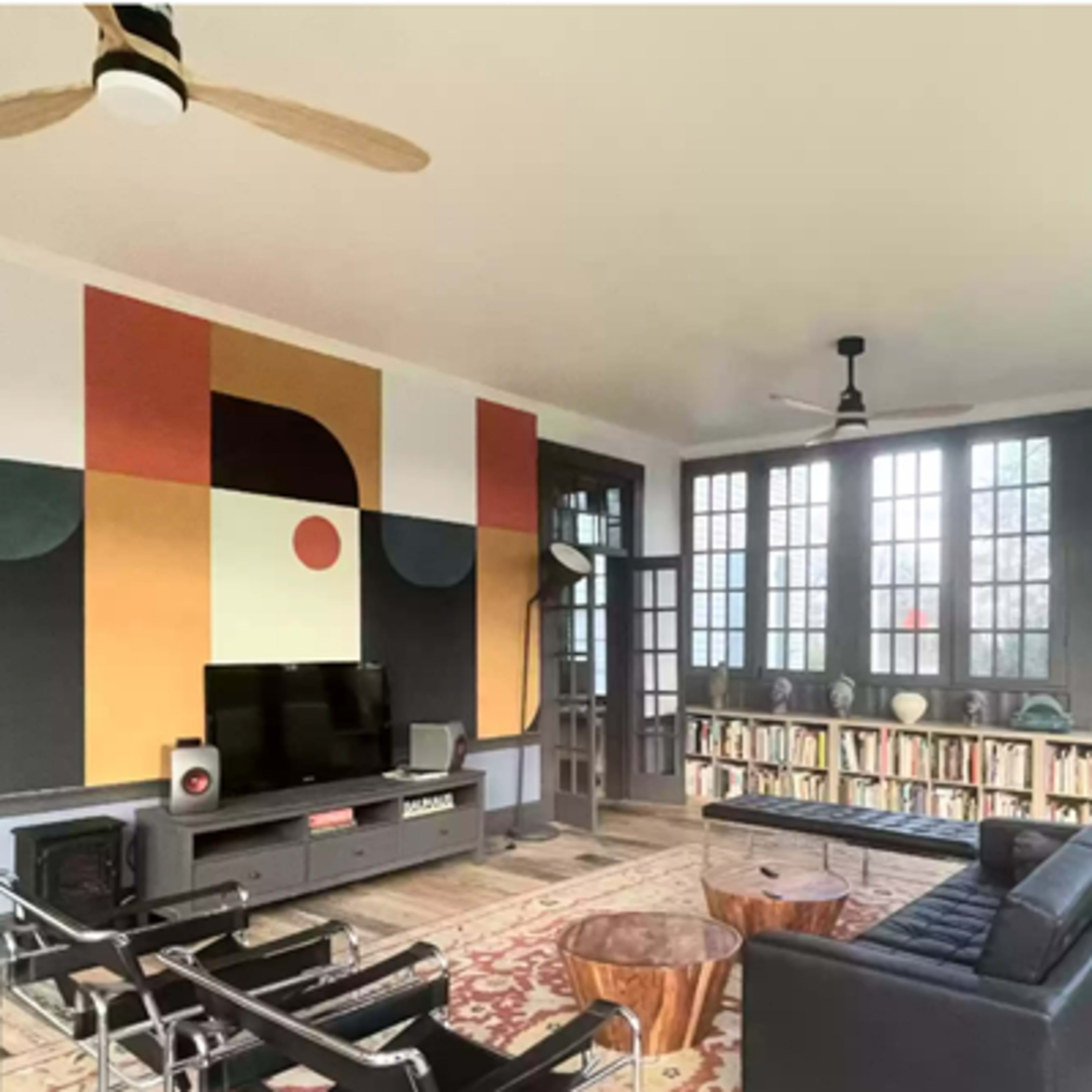A modern living room with large windows, featuring a black sofa, a wooden coffee table, a TV on an entertainment console, and a bookshelf against the wall, all set against a colorful abstract mural.