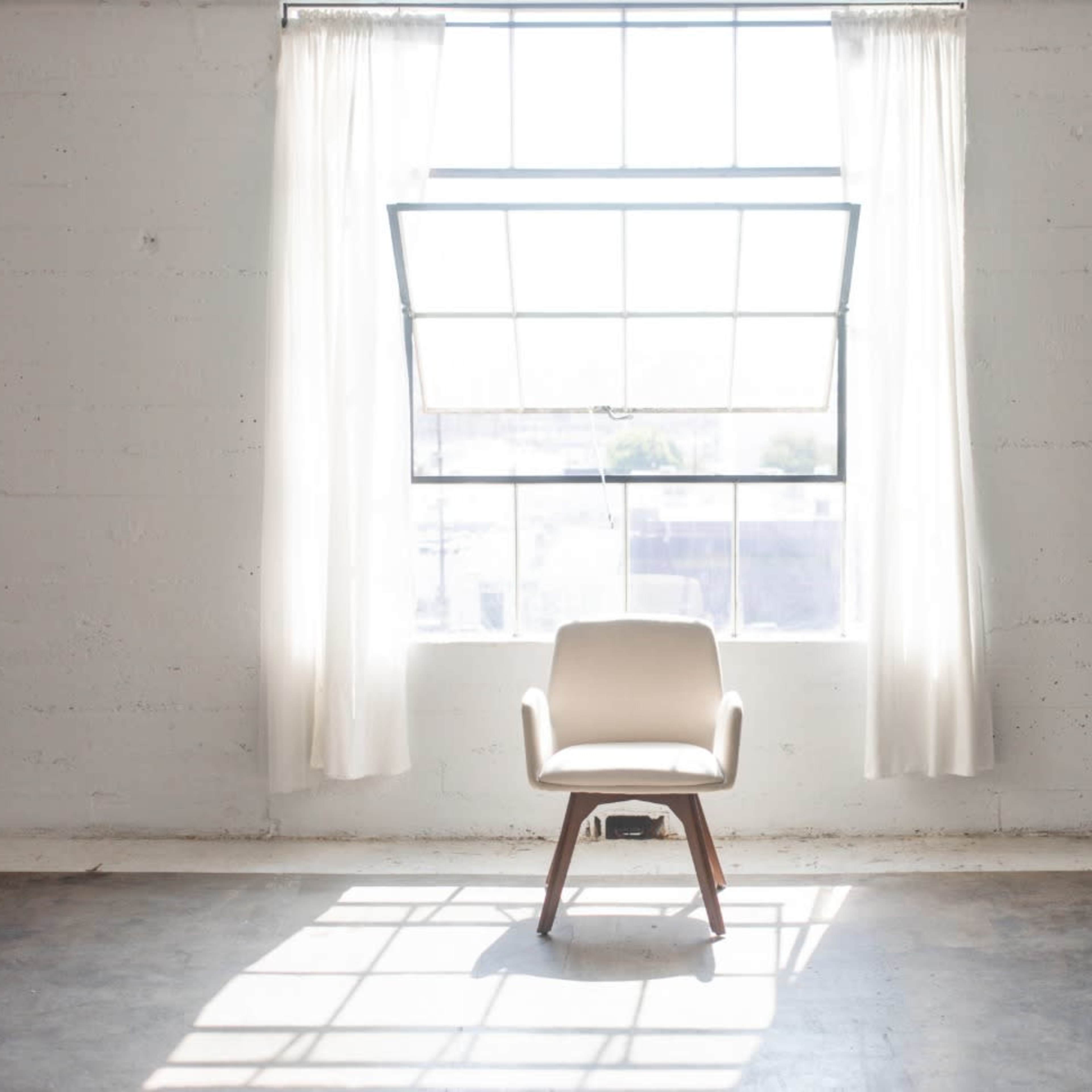 A single chair is placed beneath a large window with sheer curtains in a bright, empty room.