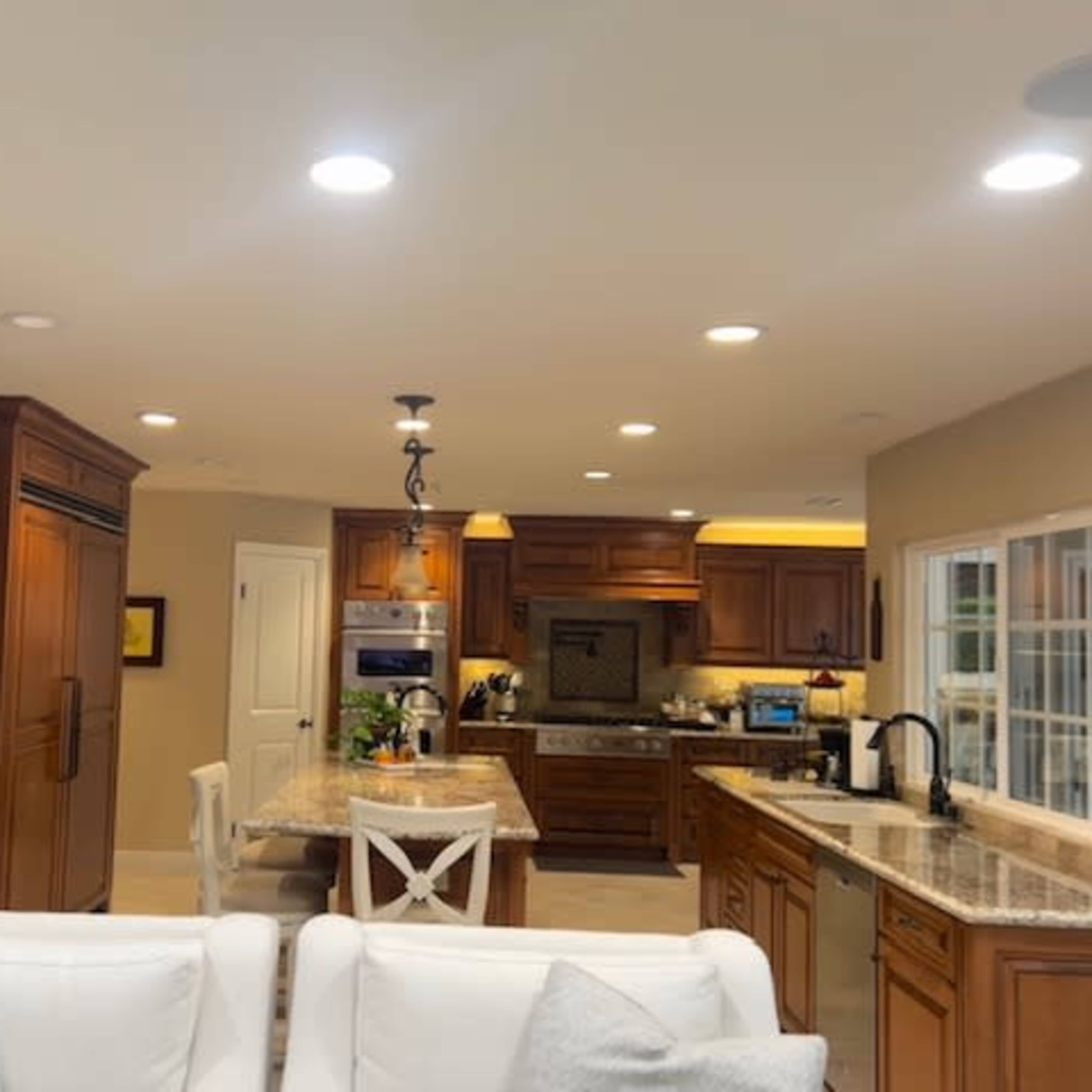 A spacious kitchen with wooden cabinetry, granite countertops, and modern appliances, viewed from a seating area with white sofas.