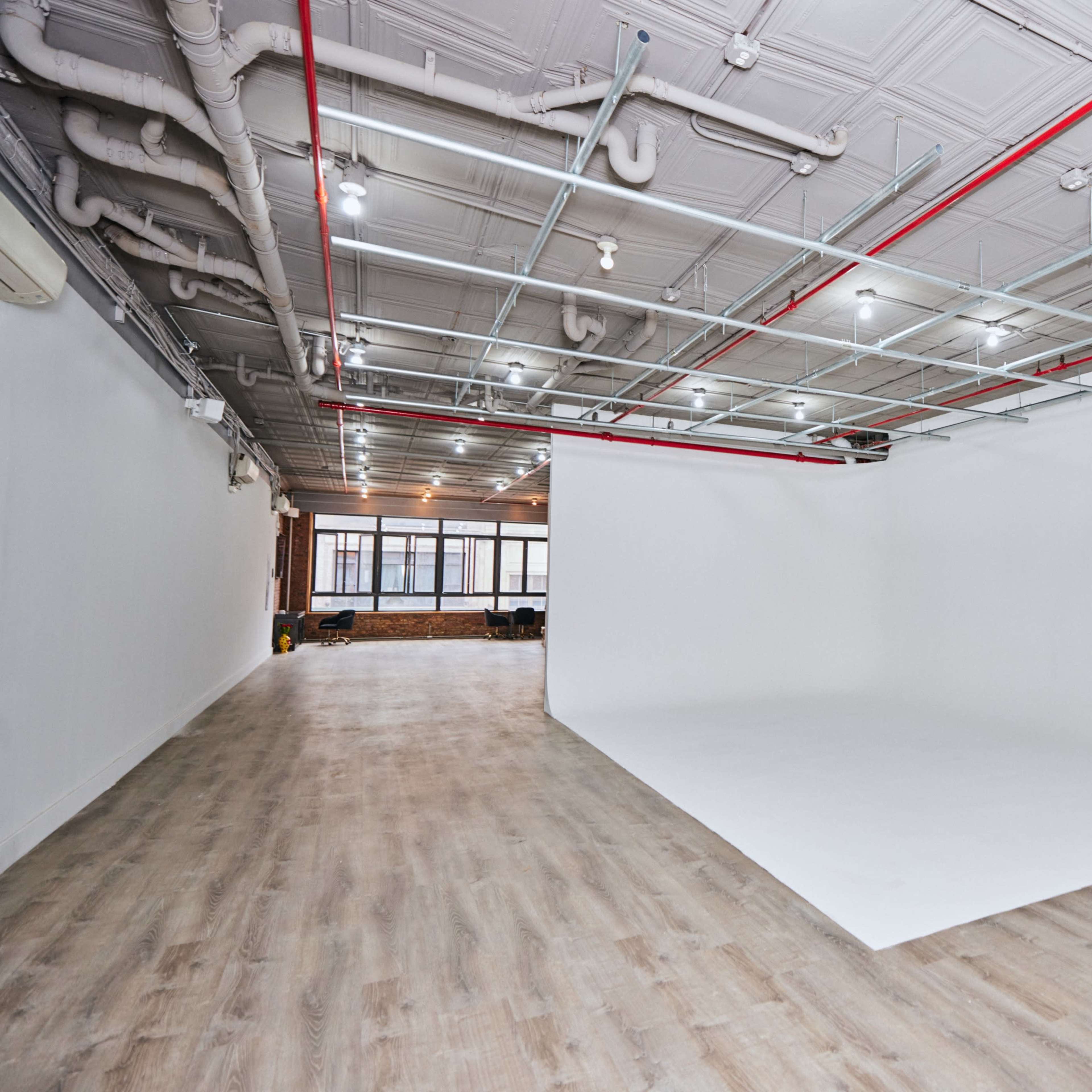 A spacious studio with a white backdrop, wooden flooring, and overhead lighting along with exposed pipes and a rolling stand.