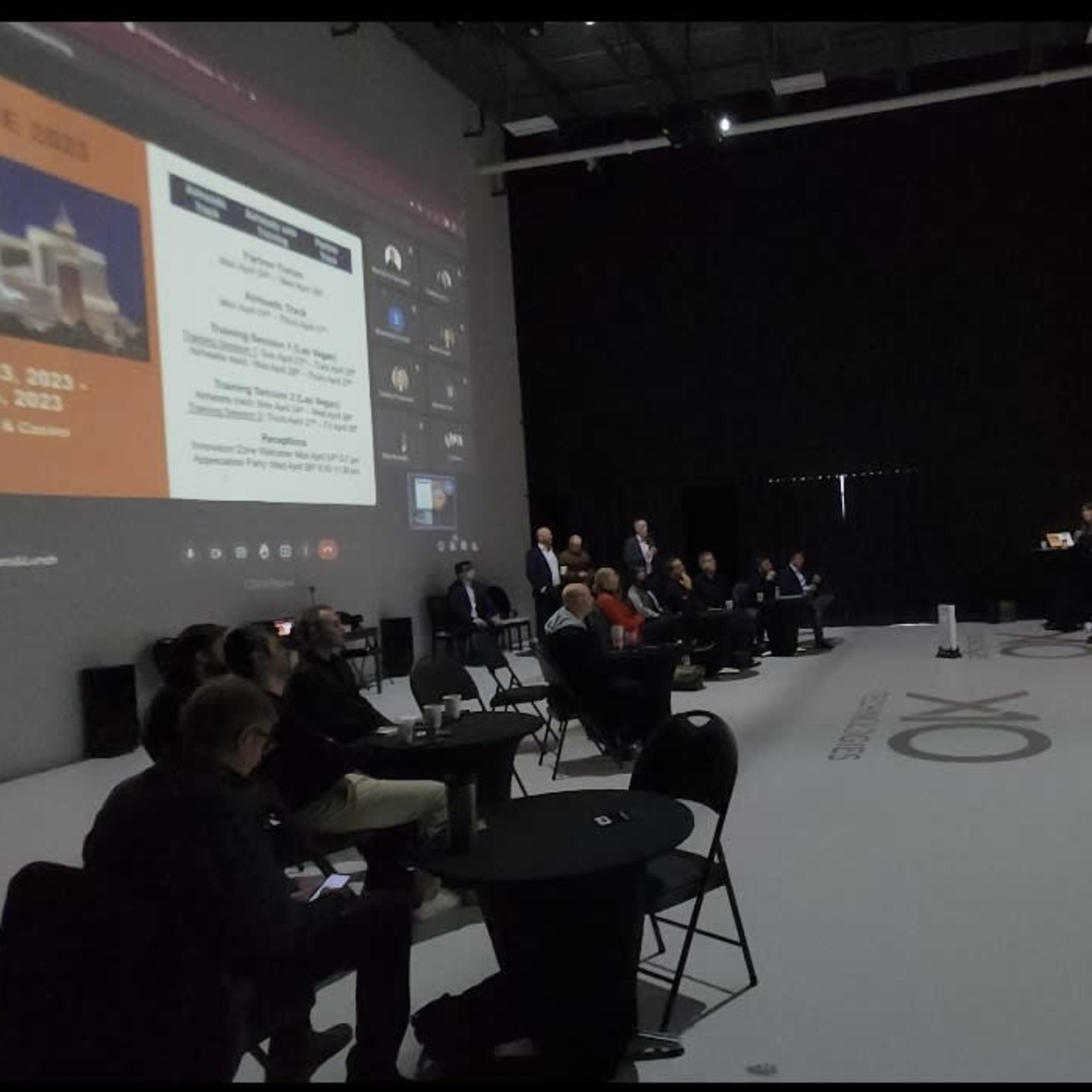 A presentation is taking place in a large, dimly lit room with several attendees seated at tables, while a speaker stands in front of a large screen displaying event information.