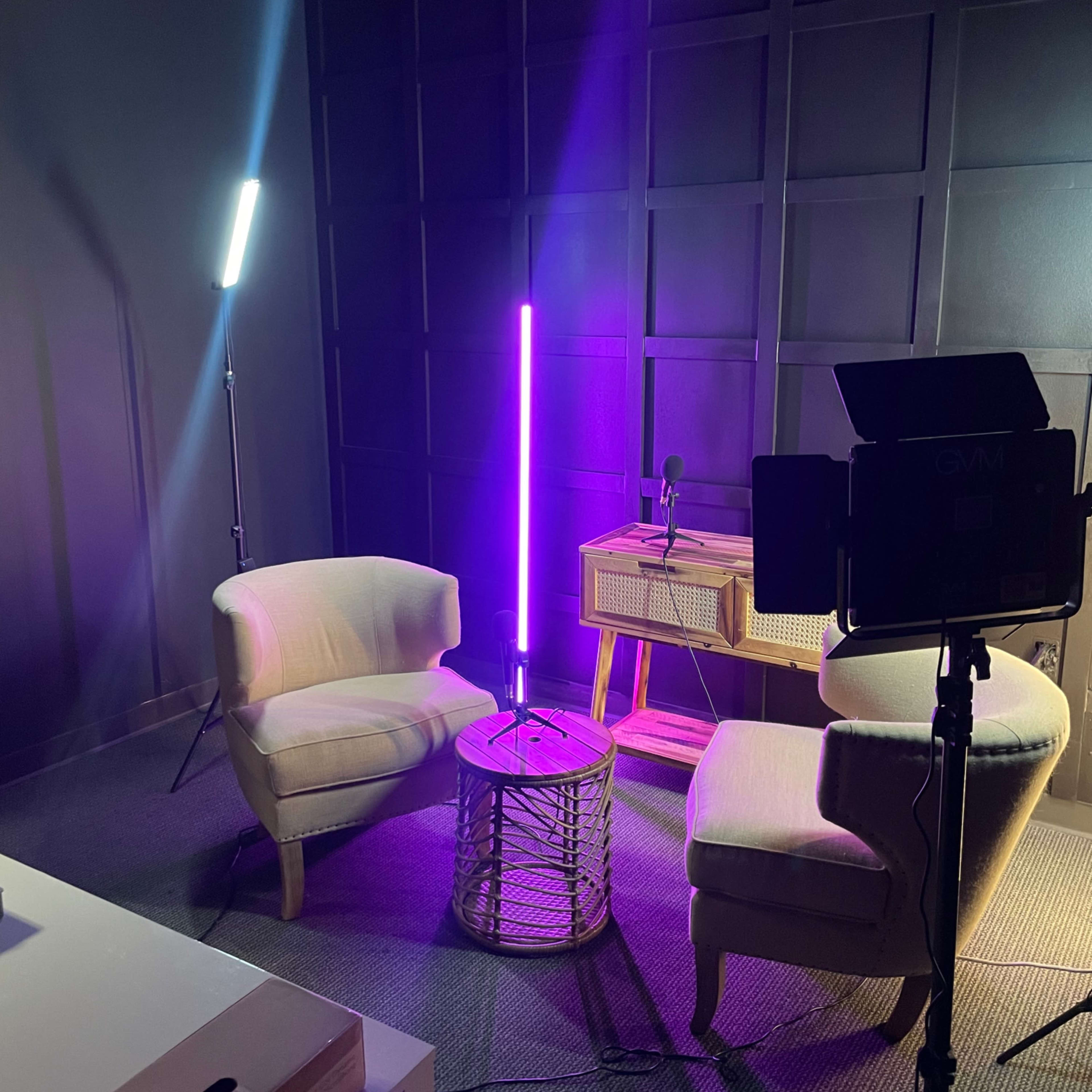 A small studio setup with two light sources, two chairs, a table made of woven material, and a microphone on a stand, all against a dark wall.
