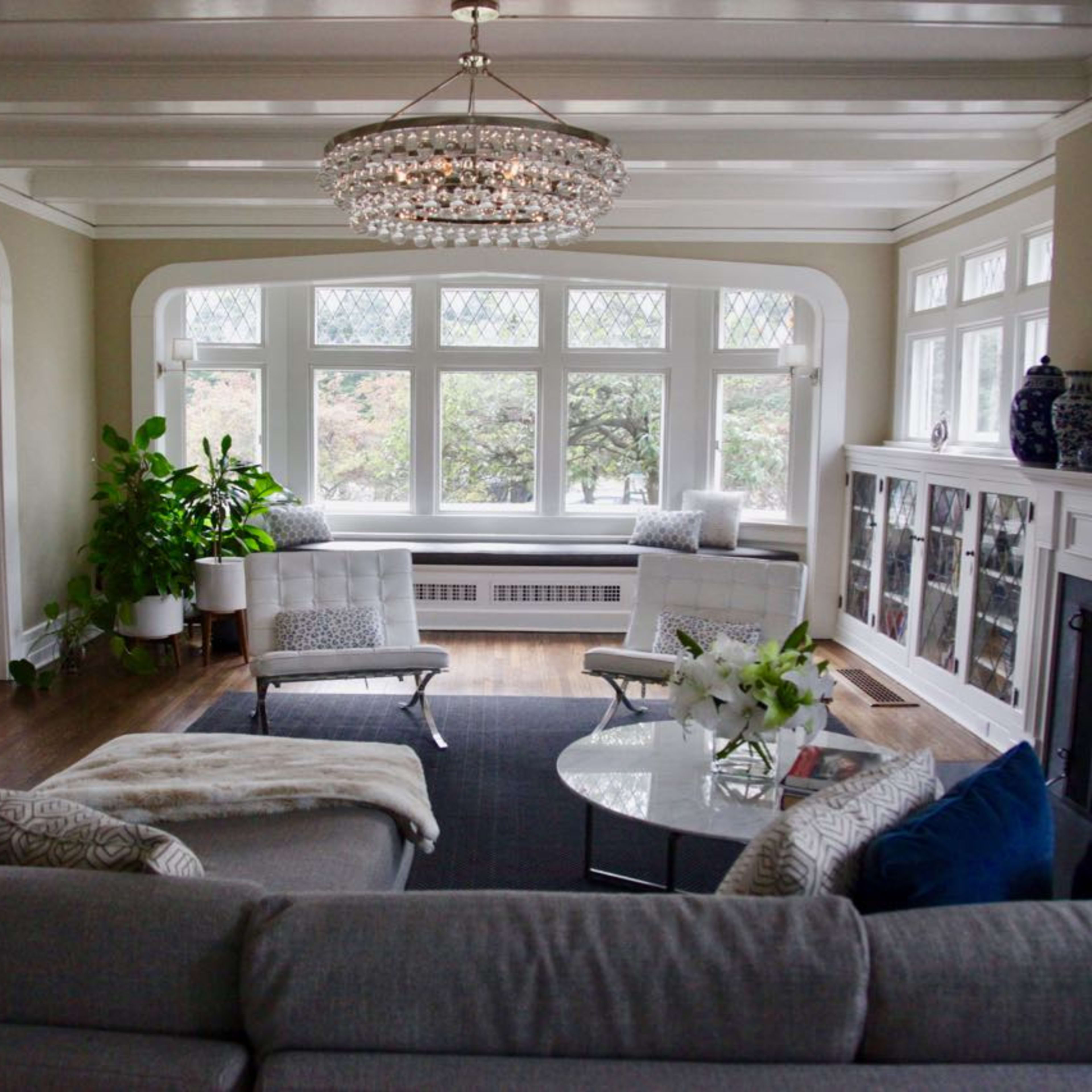 A modern living room with large windows, a crystal chandelier, and a combination of seating options arranged around a circular coffee table.