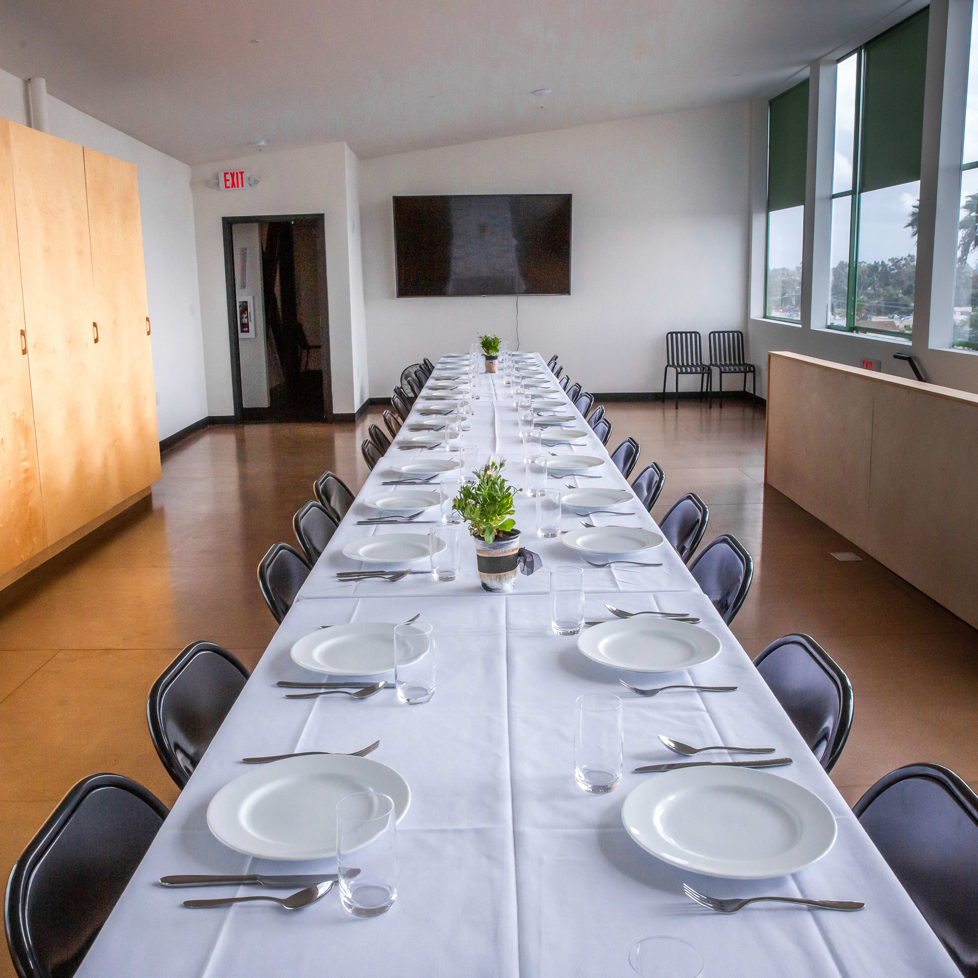 A long banquet table set with white plates and utensils is arranged in a brightly lit room with large windows and minimalist decor.