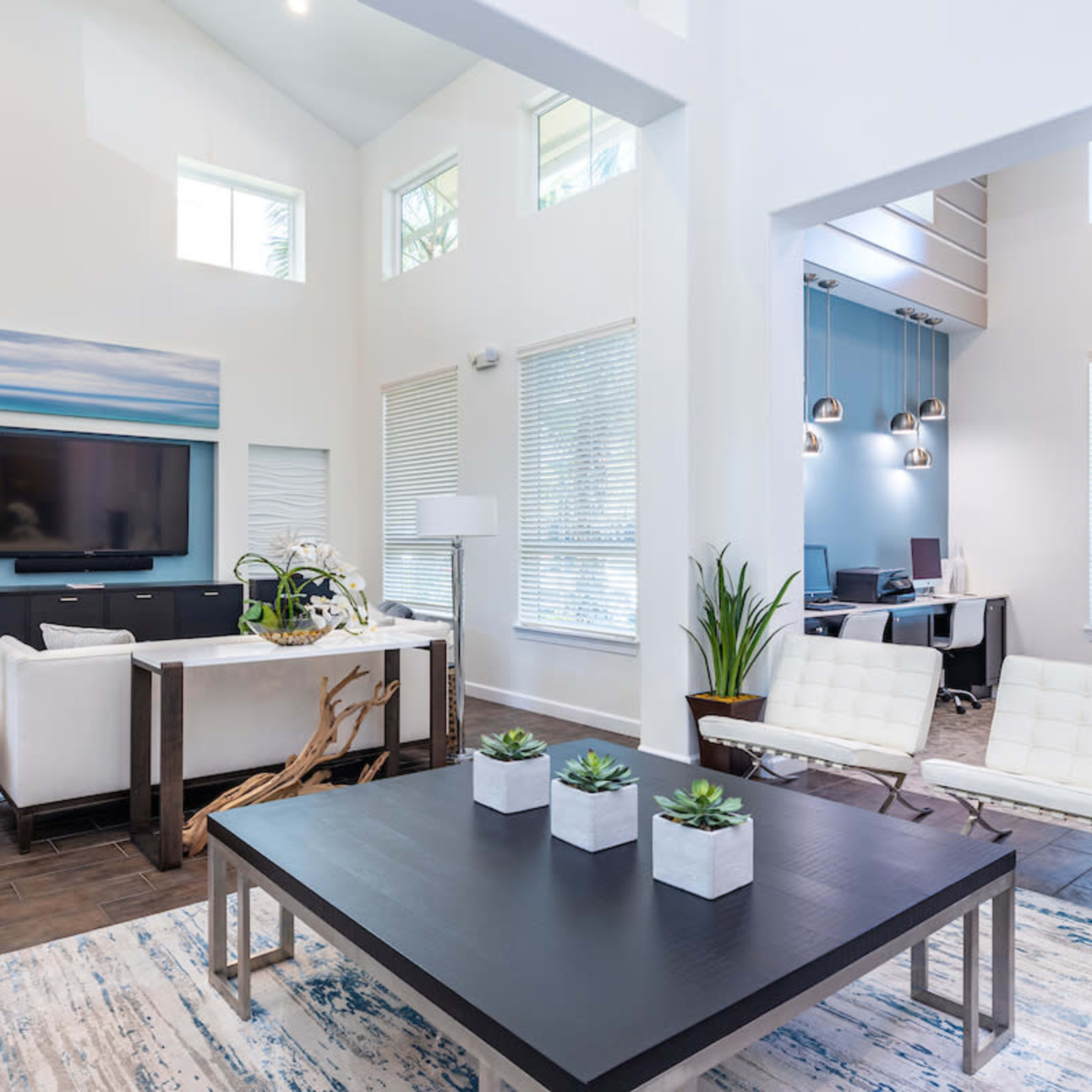A modern living room with a white couch, a large coffee table, a television mounted on the wall, and a workspace area visible through an open archway.