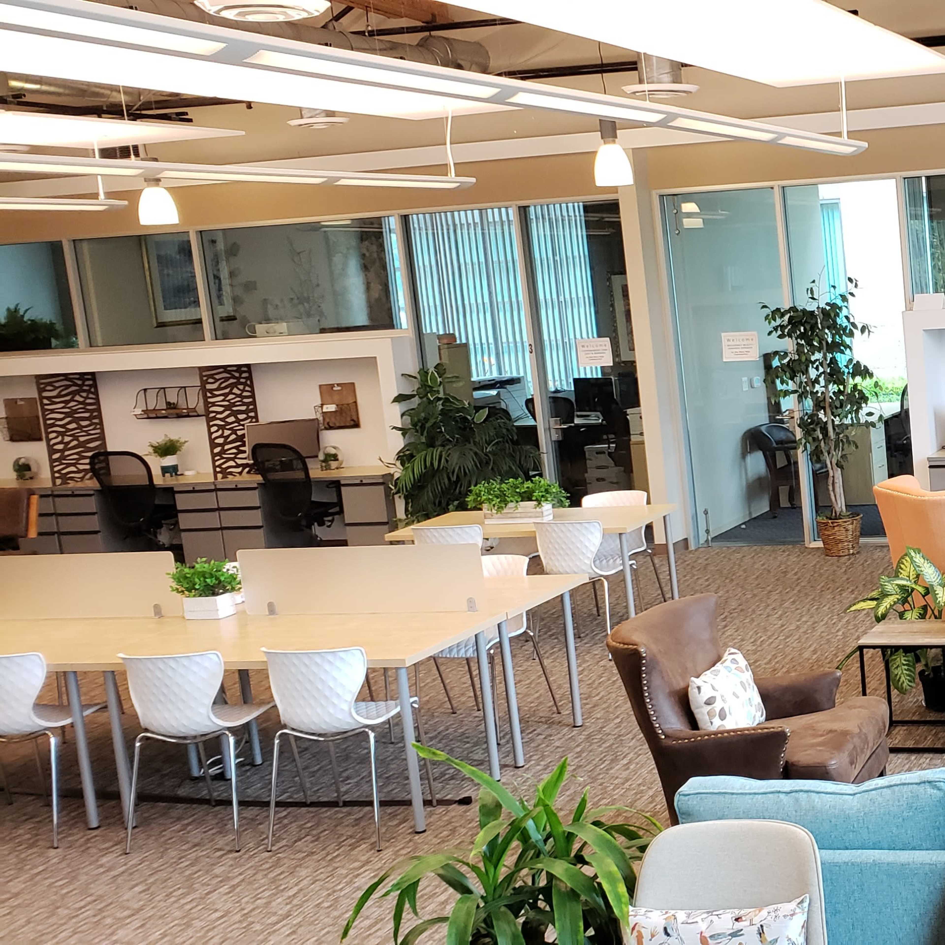 A modern office space featuring a variety of seating arrangements, greenery, and workstations organized around a large communal table.