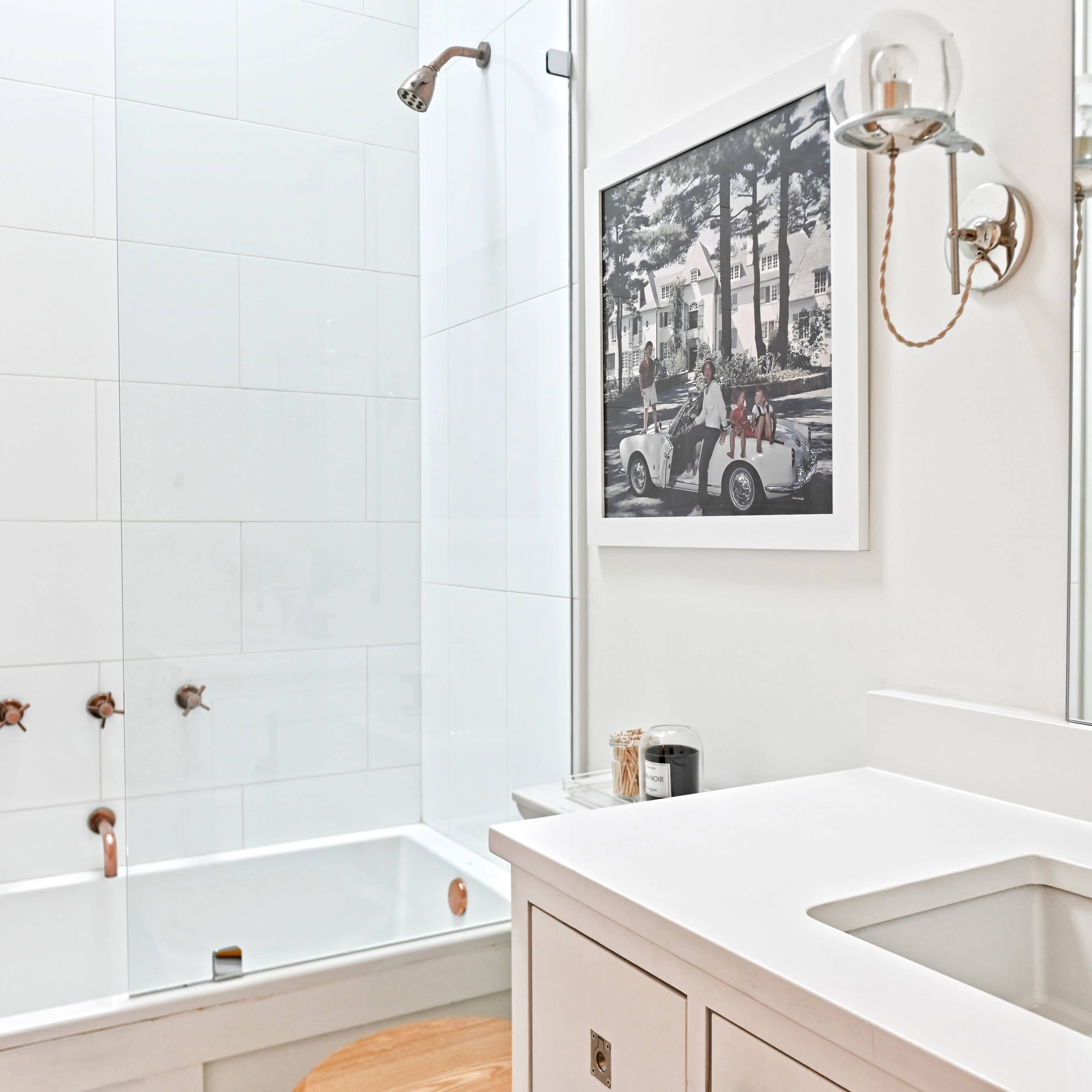 A modern bathroom with a white sink, a tub with a glass shower enclosure, and a framed black-and-white photograph on the wall.