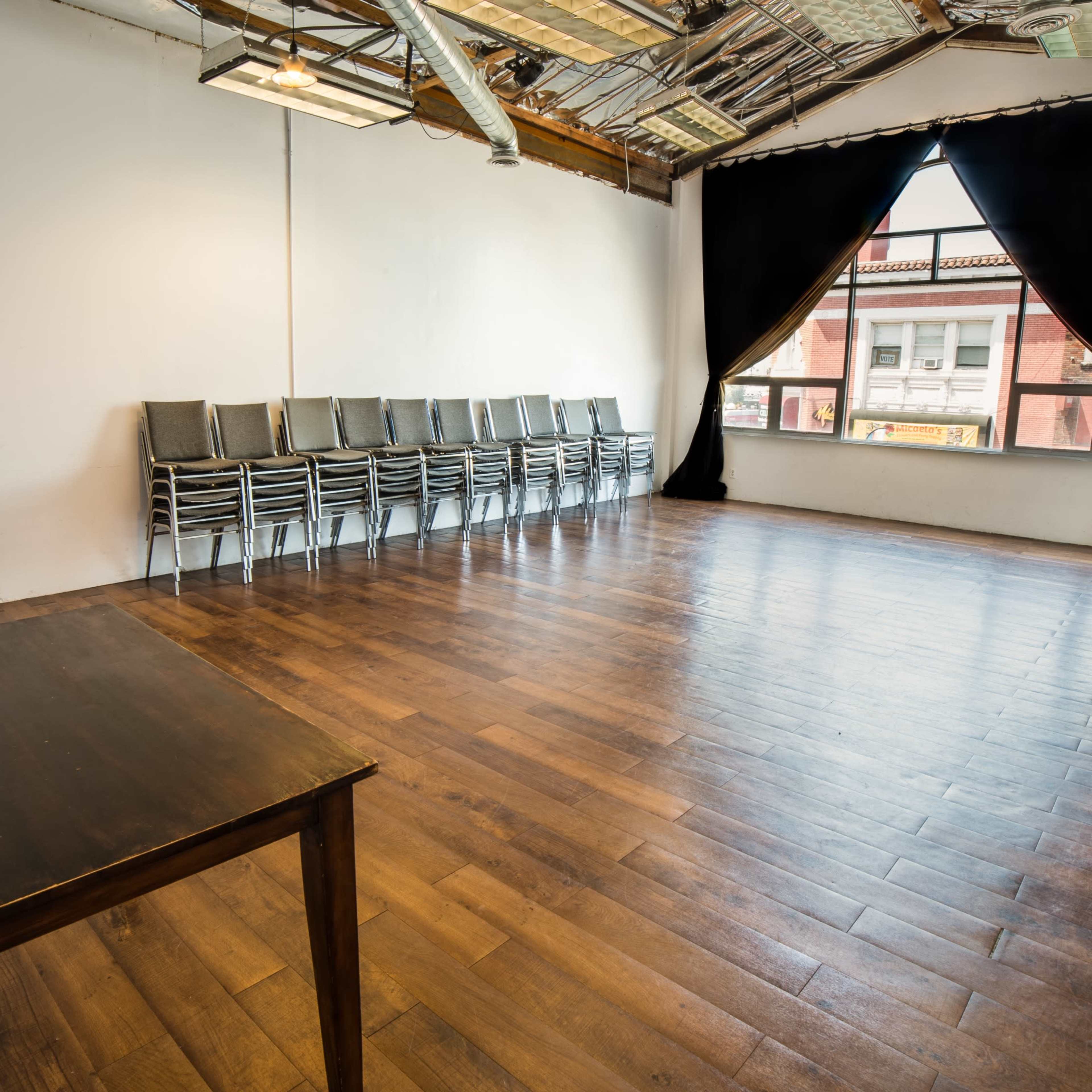 An empty room with wooden flooring, a row of stacked chairs against one wall, and a large window covered by a black curtain.