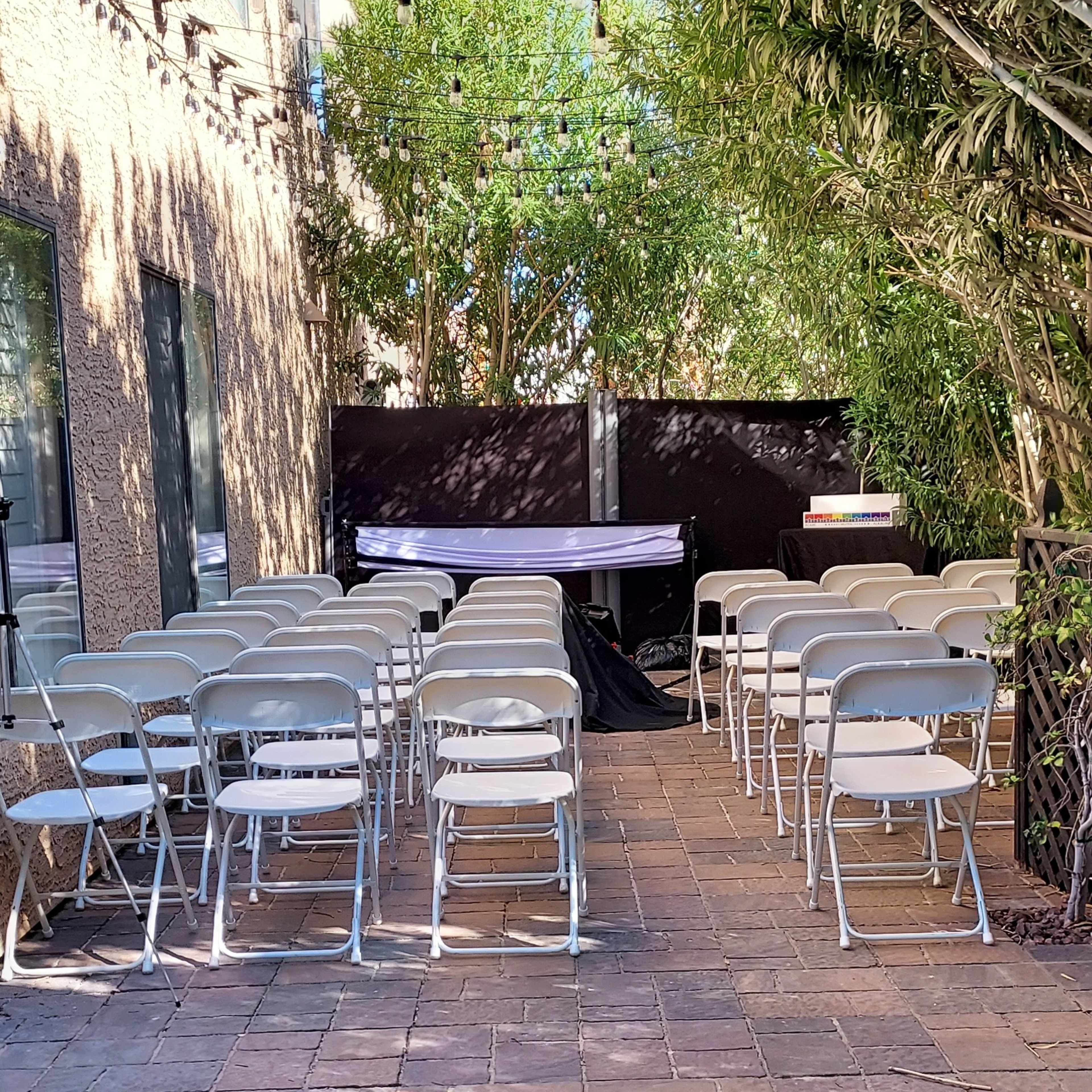 A row of folding chairs is set up on a paved area surrounded by green grass and plants, with a backdrop for an event in a courtyard.