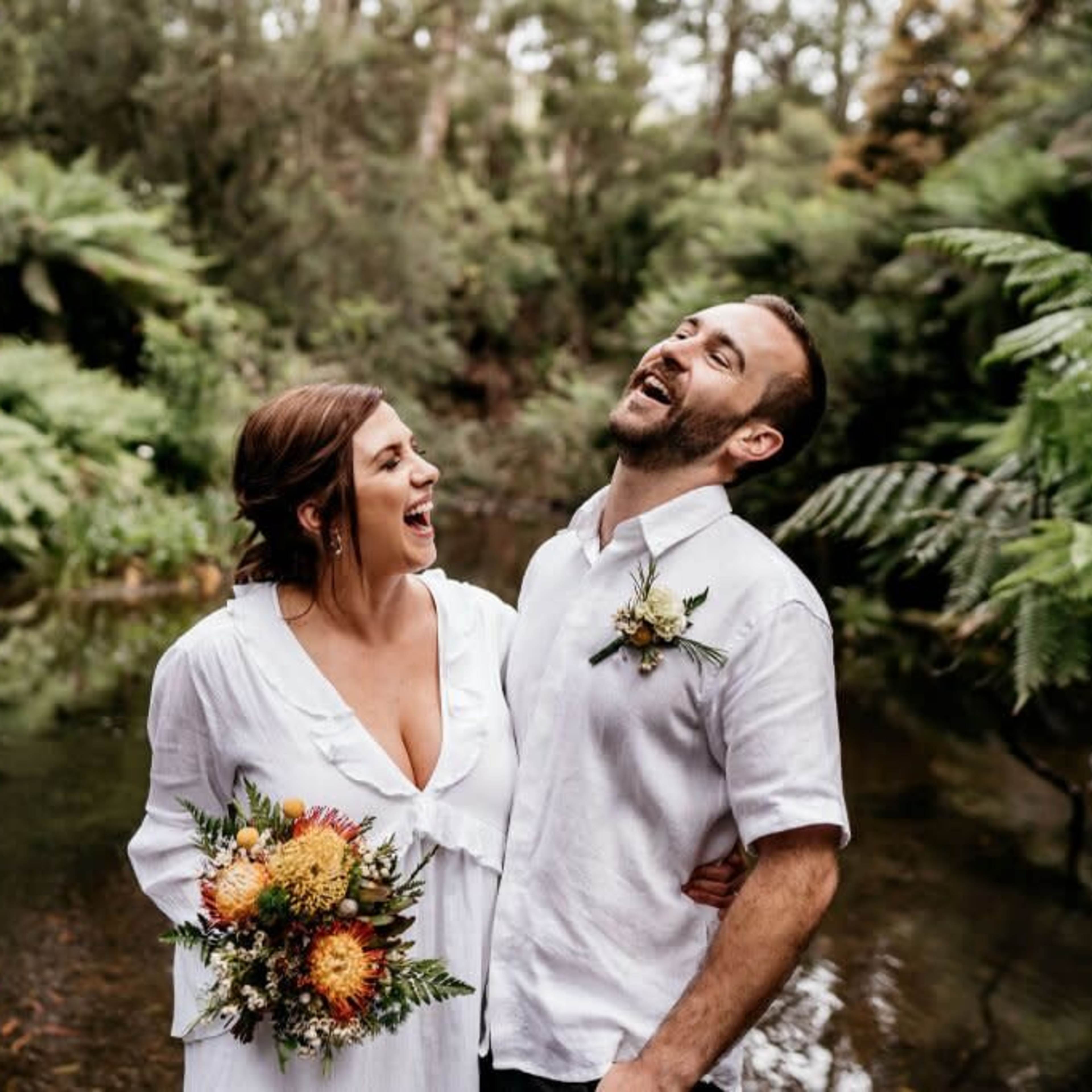 A couple smiles and laughs together while standing near a creek surrounded by lush greenery.