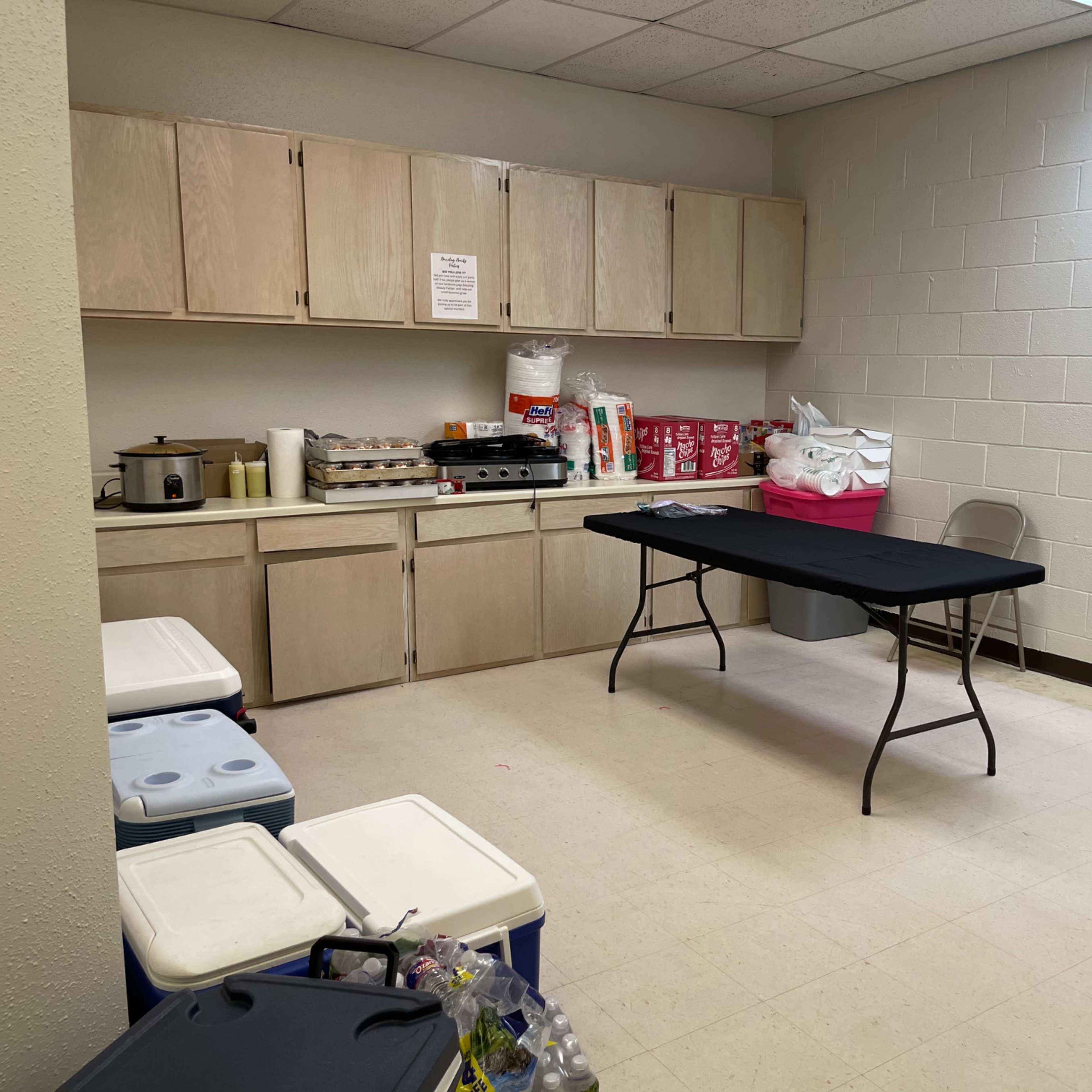 A small kitchenette features a table with a black cloth, surrounded by cabinets and coolers, with various food items and supplies on the counters.