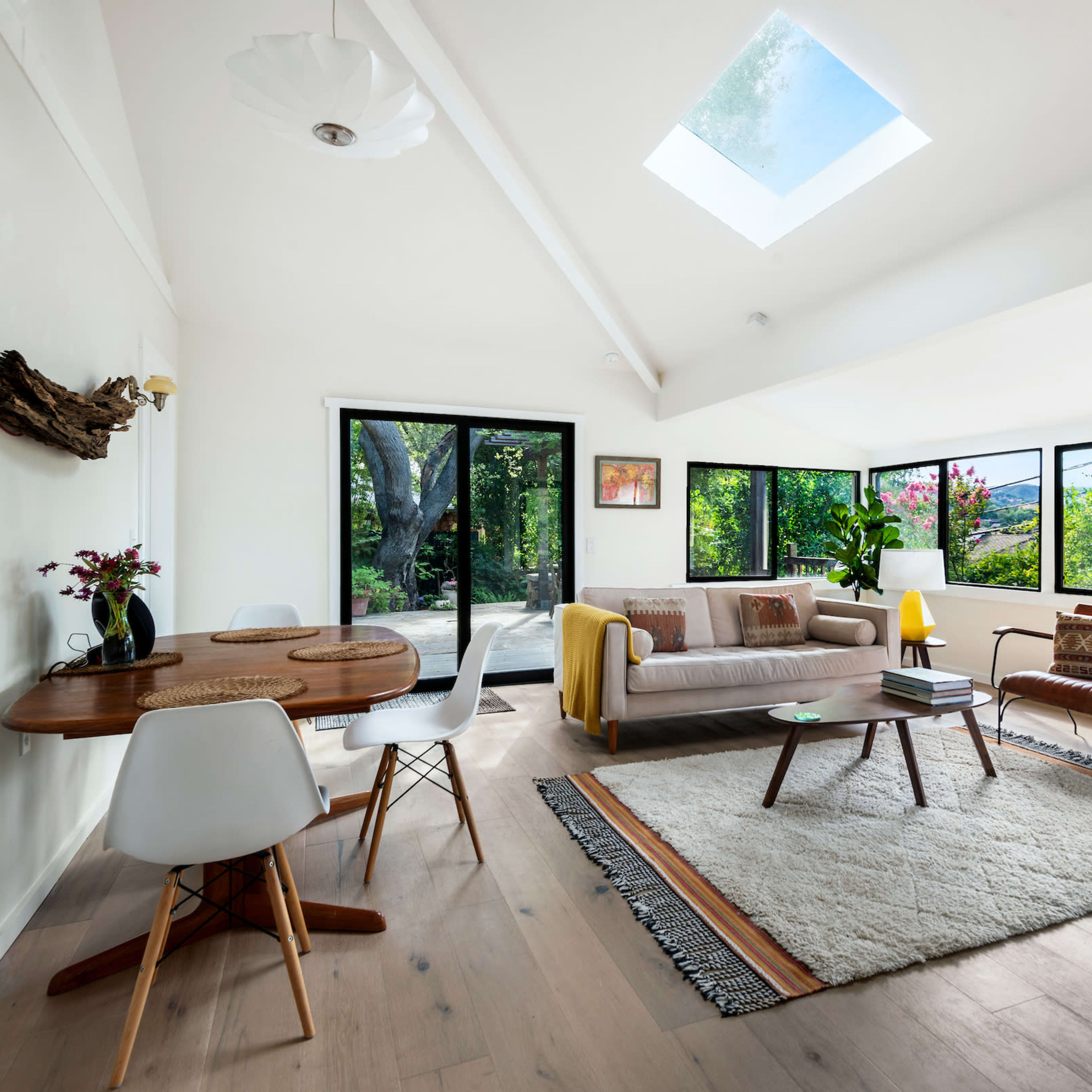 A bright living room features large windows overlooking greenery, with a sofa, dining table, and a patterned rug on a wooden floor.