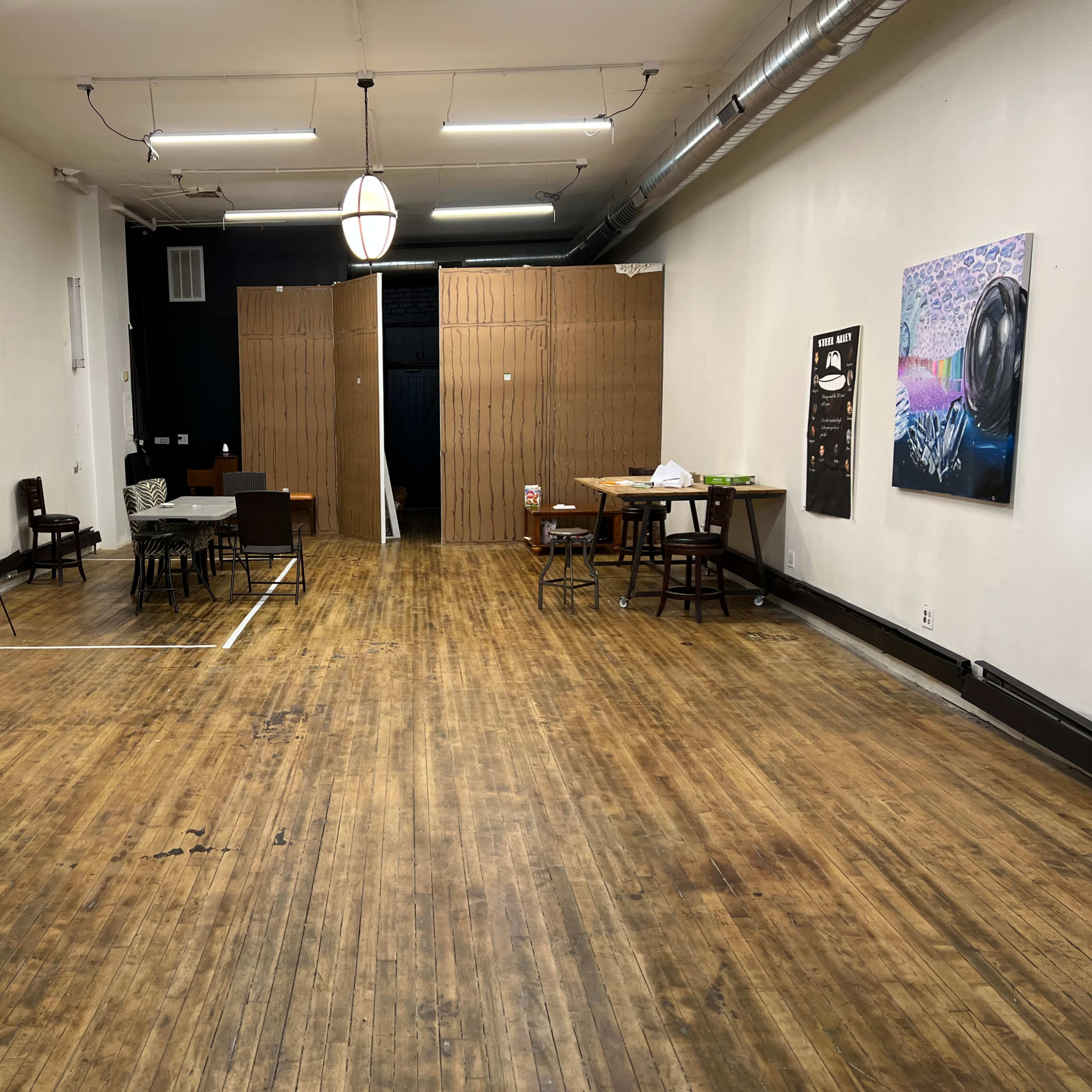 A spacious, empty room with wooden floors, featuring a table and chairs set up on one side, and a black wall with a wooden partition in the background.