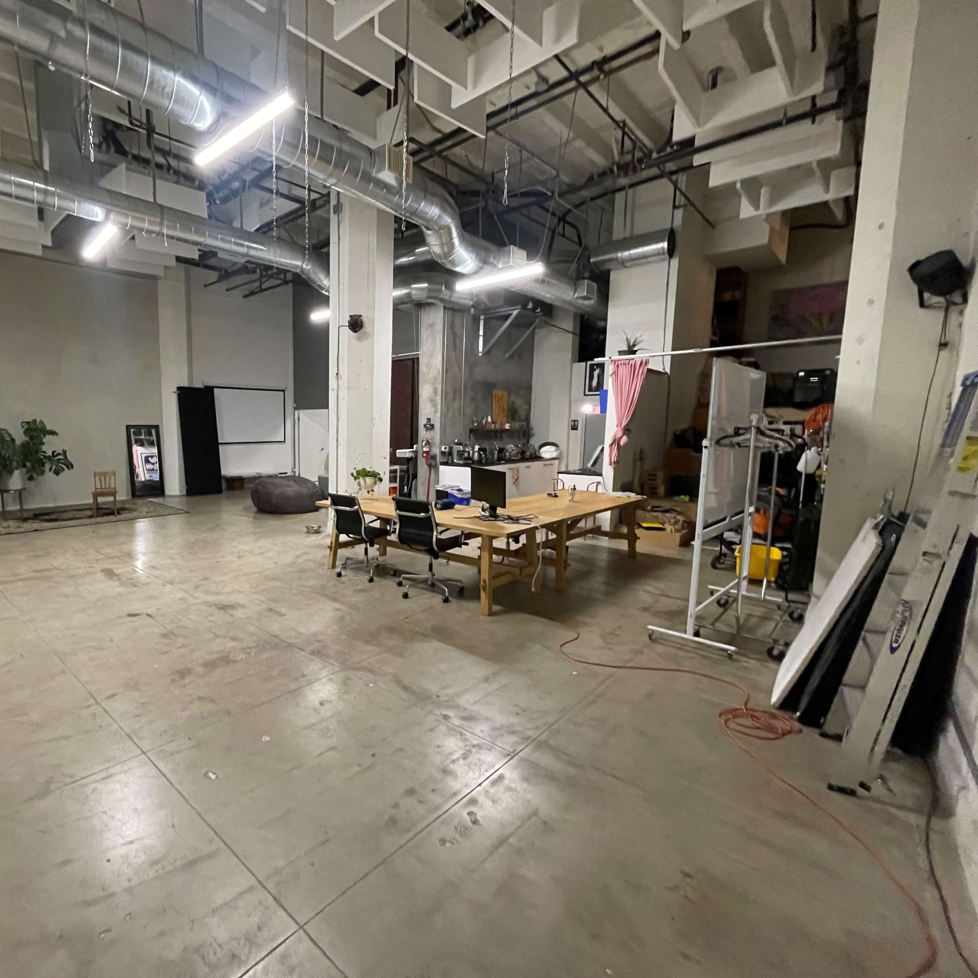 A spacious, empty room with concrete flooring, equipped with several lights, a table surrounded by chairs, and various equipment and plants scattered around.