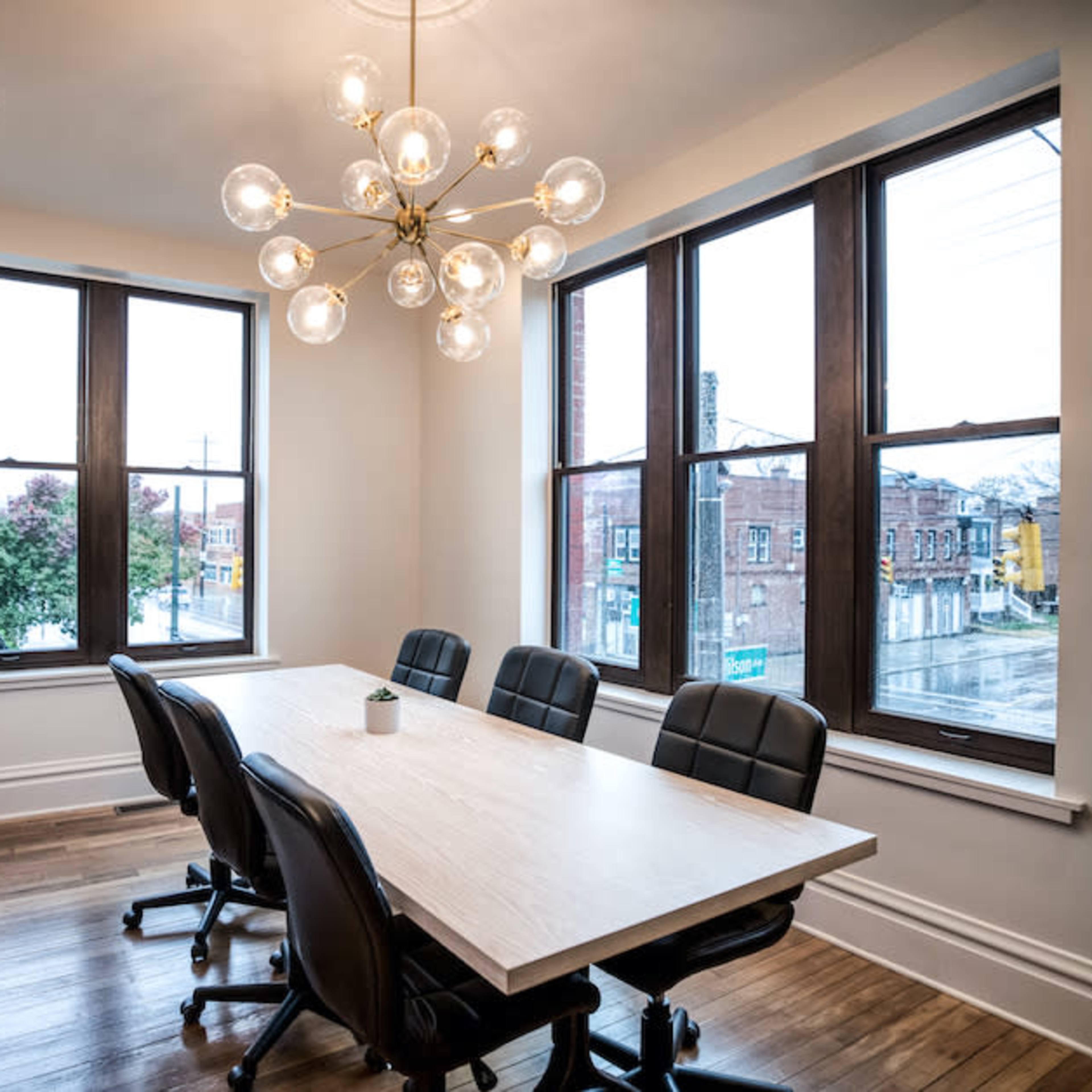 A modern conference room features a large wooden table surrounded by black chairs and is illuminated by a statement chandelier, with windows offering a view of the street outside.