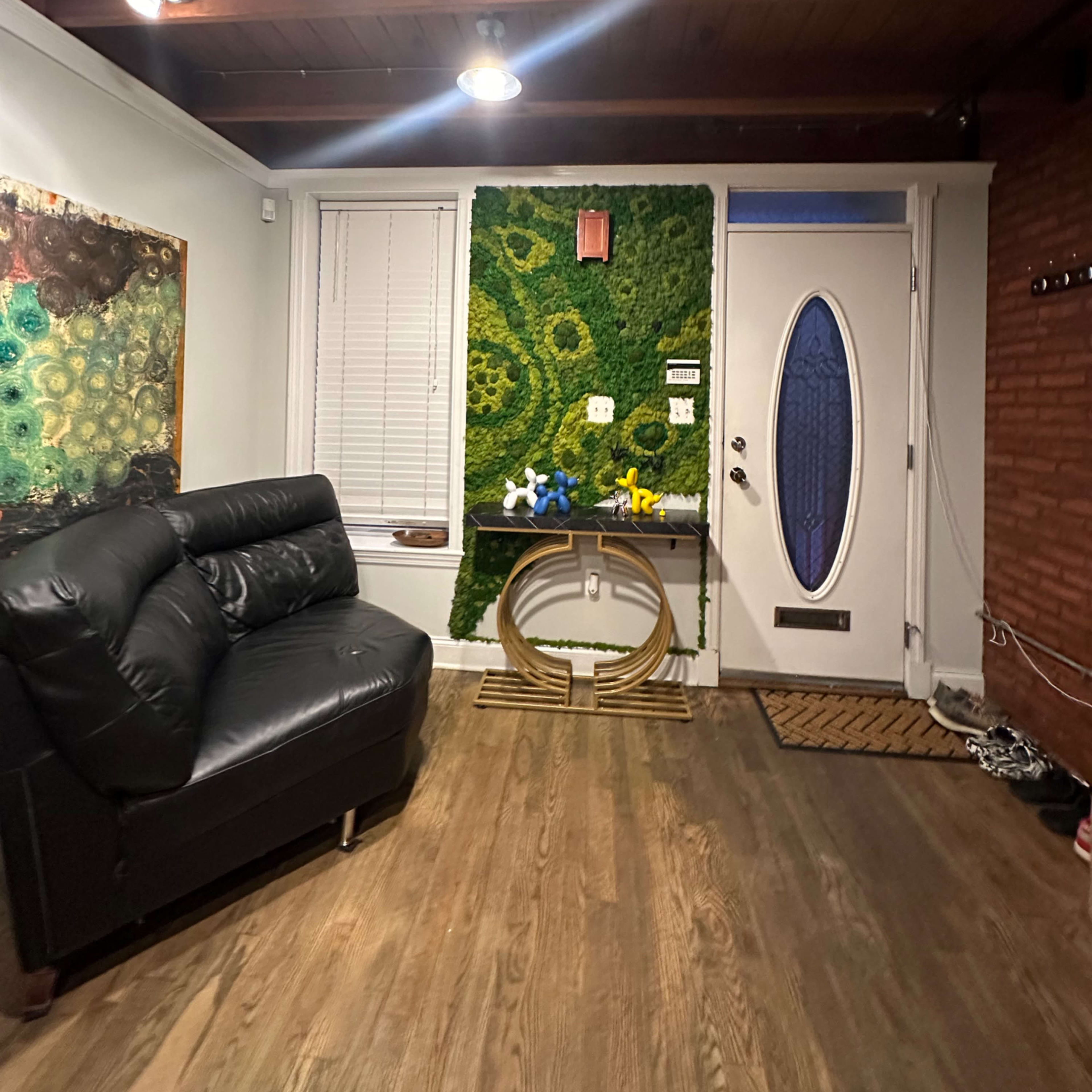 A living room with a black leather sofa, a decorative wall featuring greenery, and a door with a circular window.