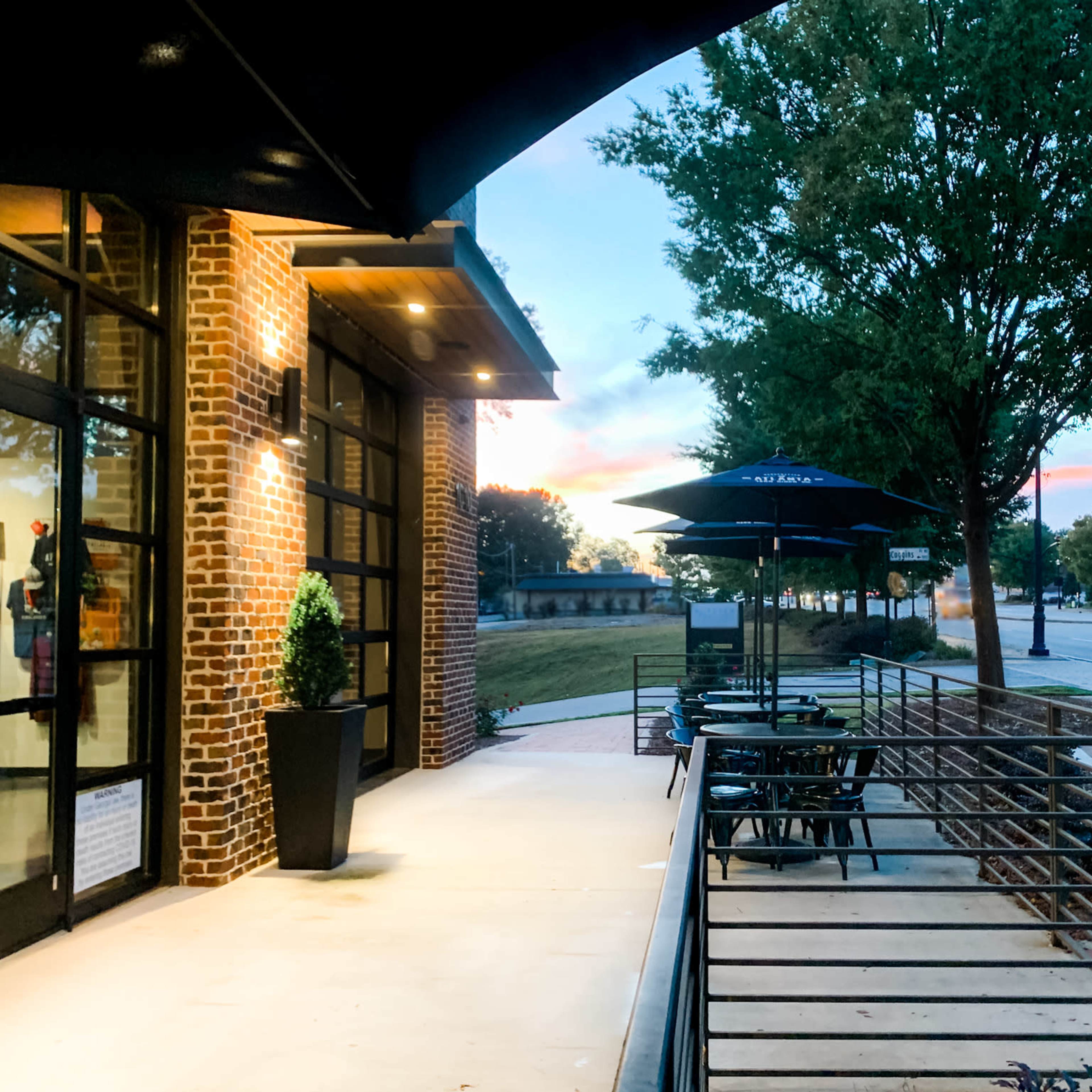 An outdoor seating area with black tables and umbrellas beside a brick building, set against a colorful sunset sky.