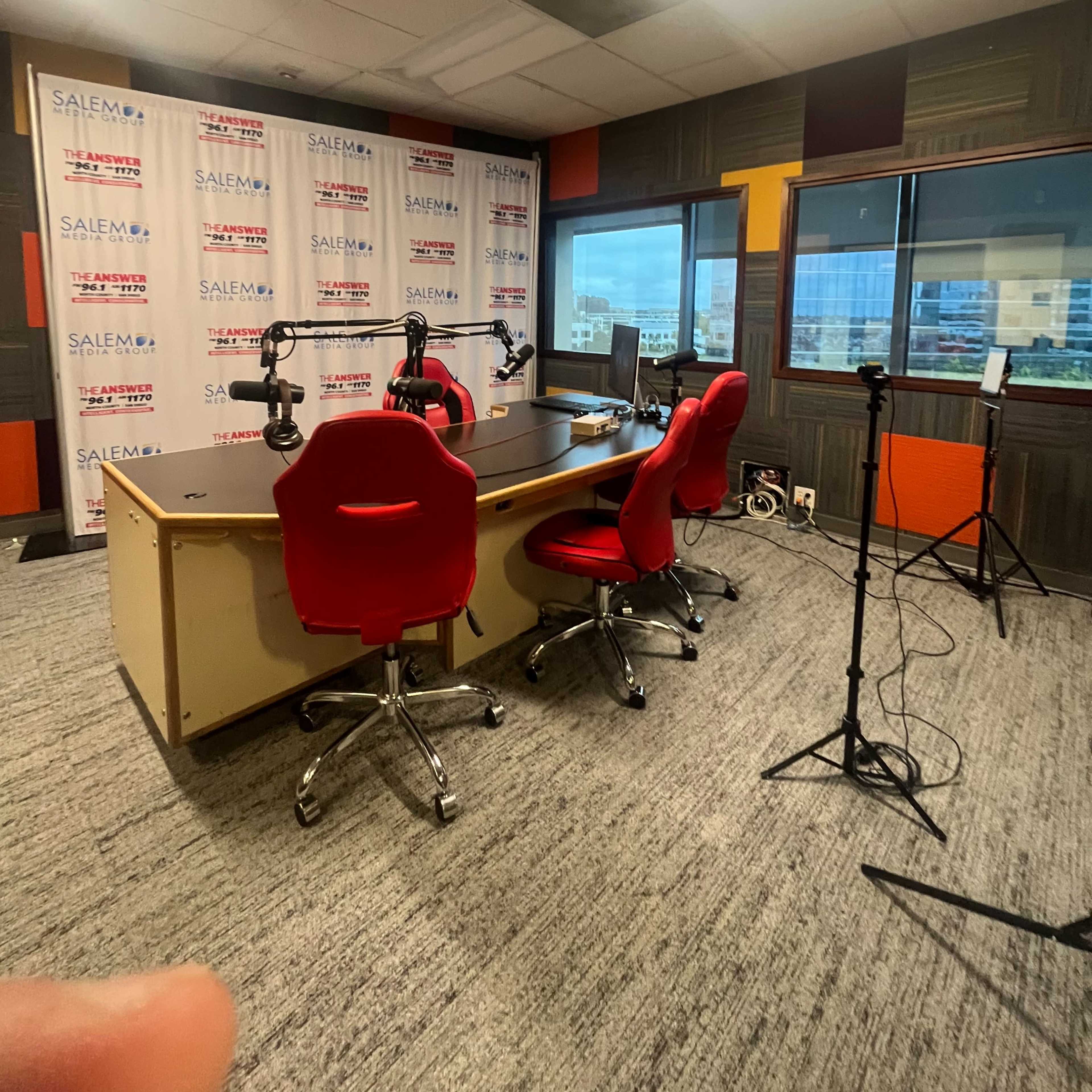 A radio studio with a table, three red chairs, and microphones set up against a backdrop of branded panels and a window overlooking a cityscape.