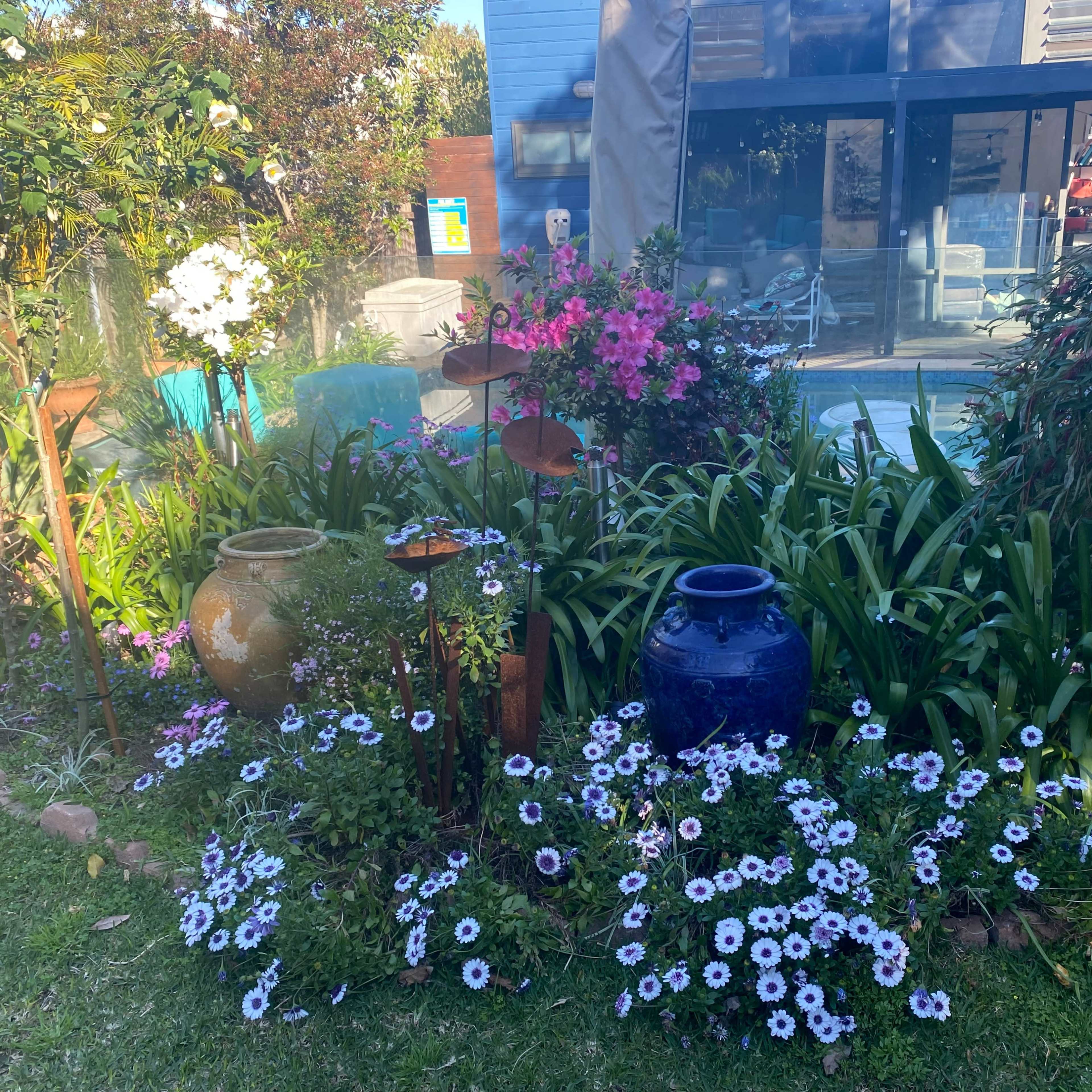 The garden features various flowers, ornamental pots, and greenery, arranged around a blue water feature.