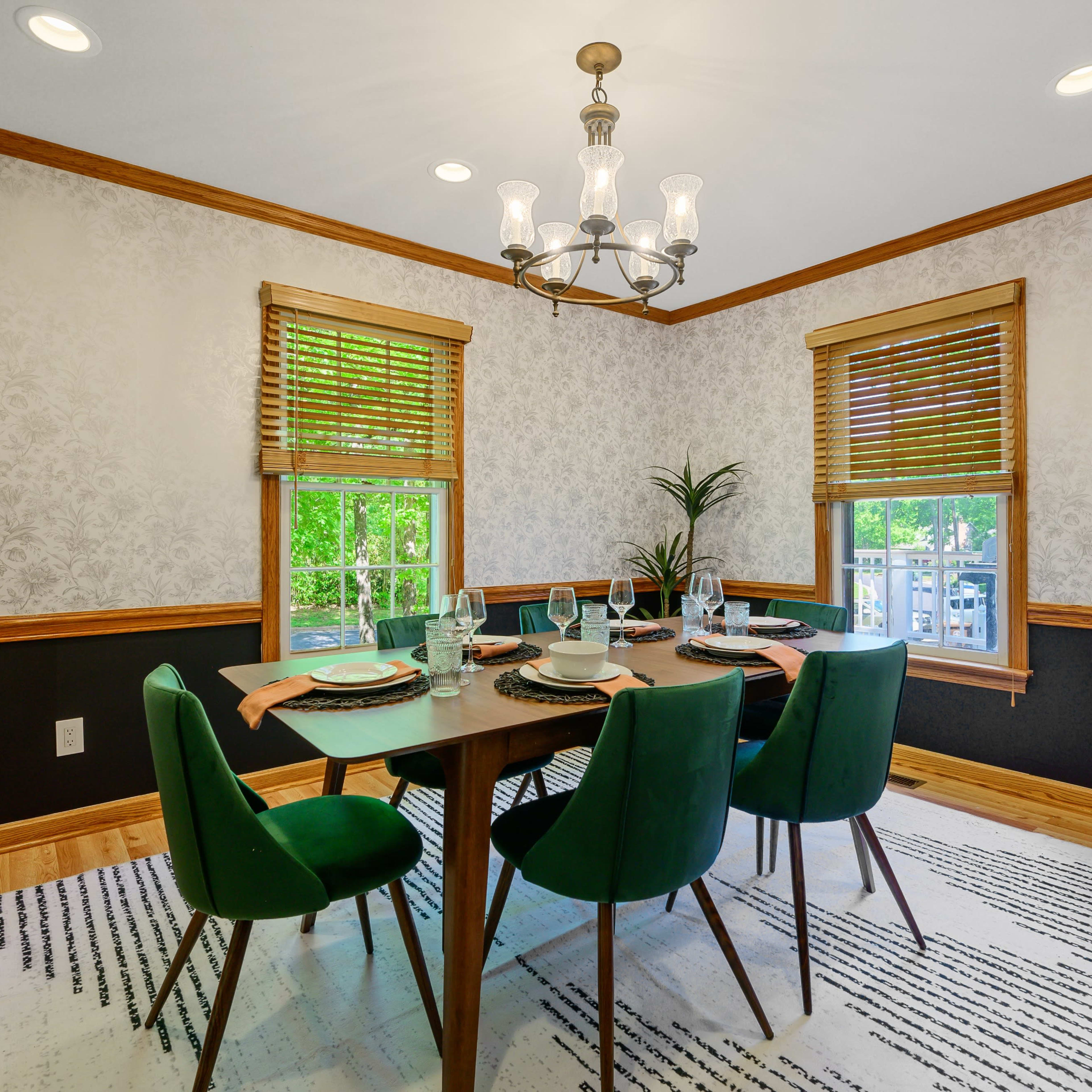 A dining room features a dark wood table surrounded by green upholstered chairs, with a chandelier overhead and patterned wallpaper.
