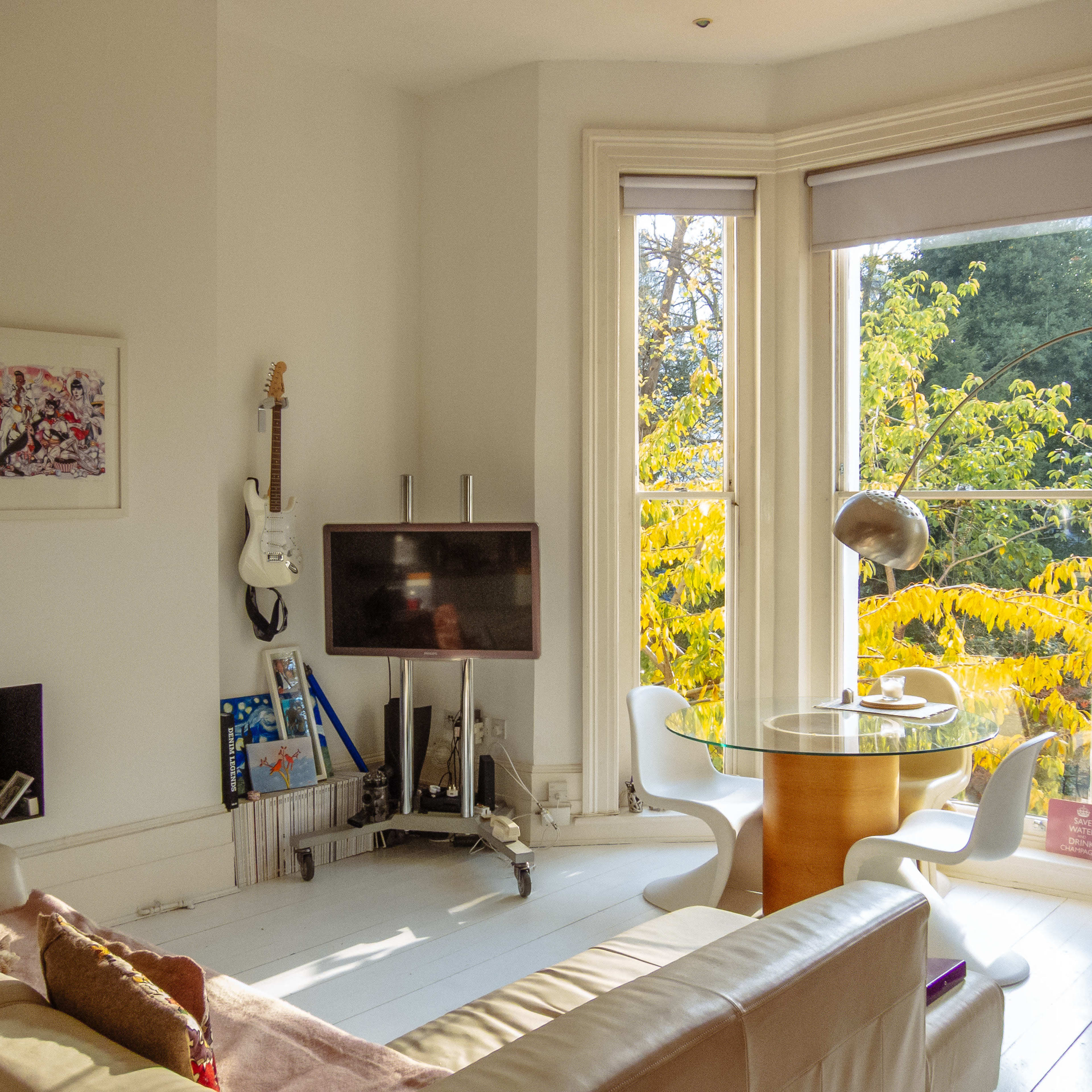A light-filled living area with a white couch, a television, a round dining table, and large windows revealing a view of trees outside.