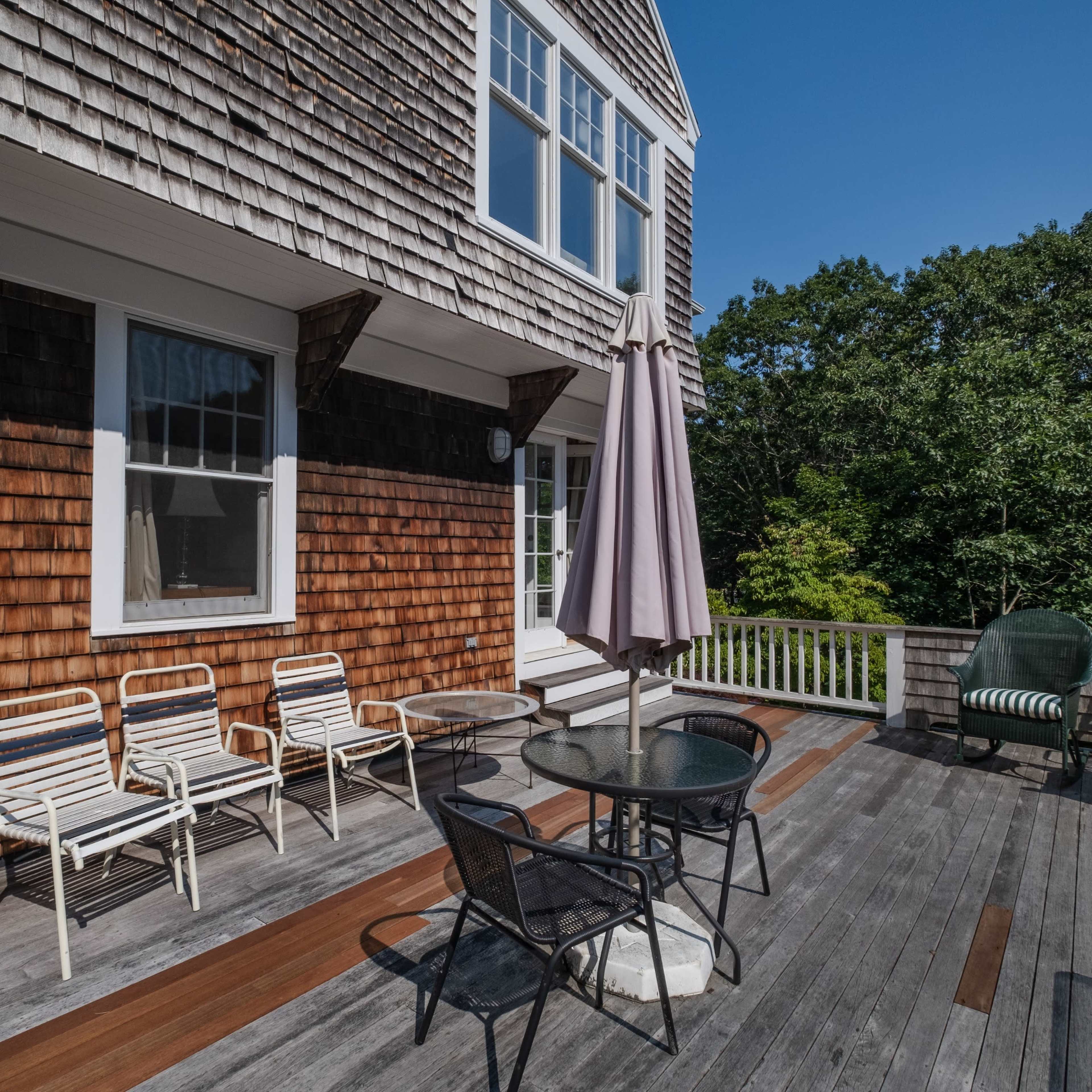A wooden deck features a table with chairs and an umbrella, surrounded by trees and a blue sky.