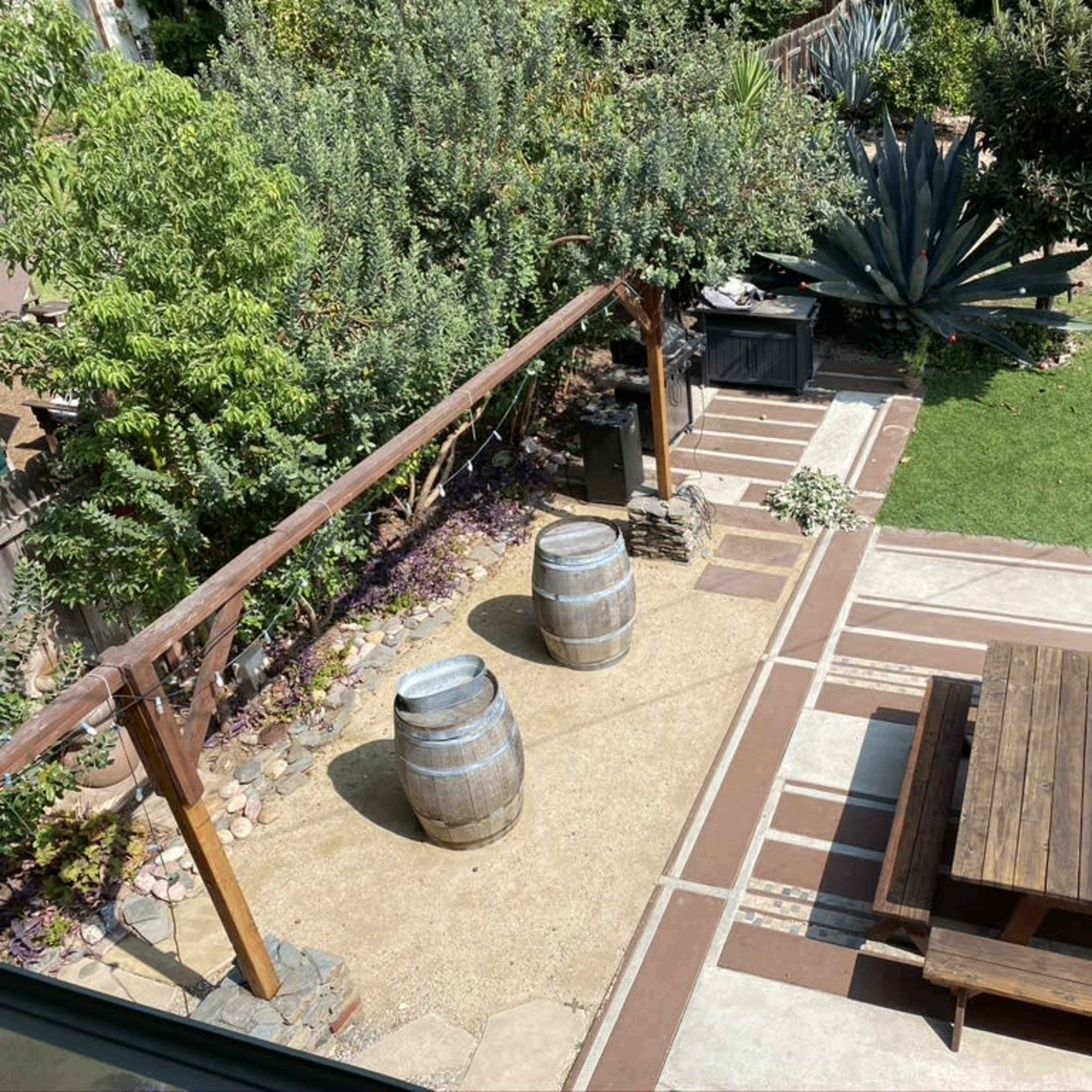 A backyard patio area featuring two wooden barrels, a picnic table, and a barbecue grill surrounded by lush greenery.