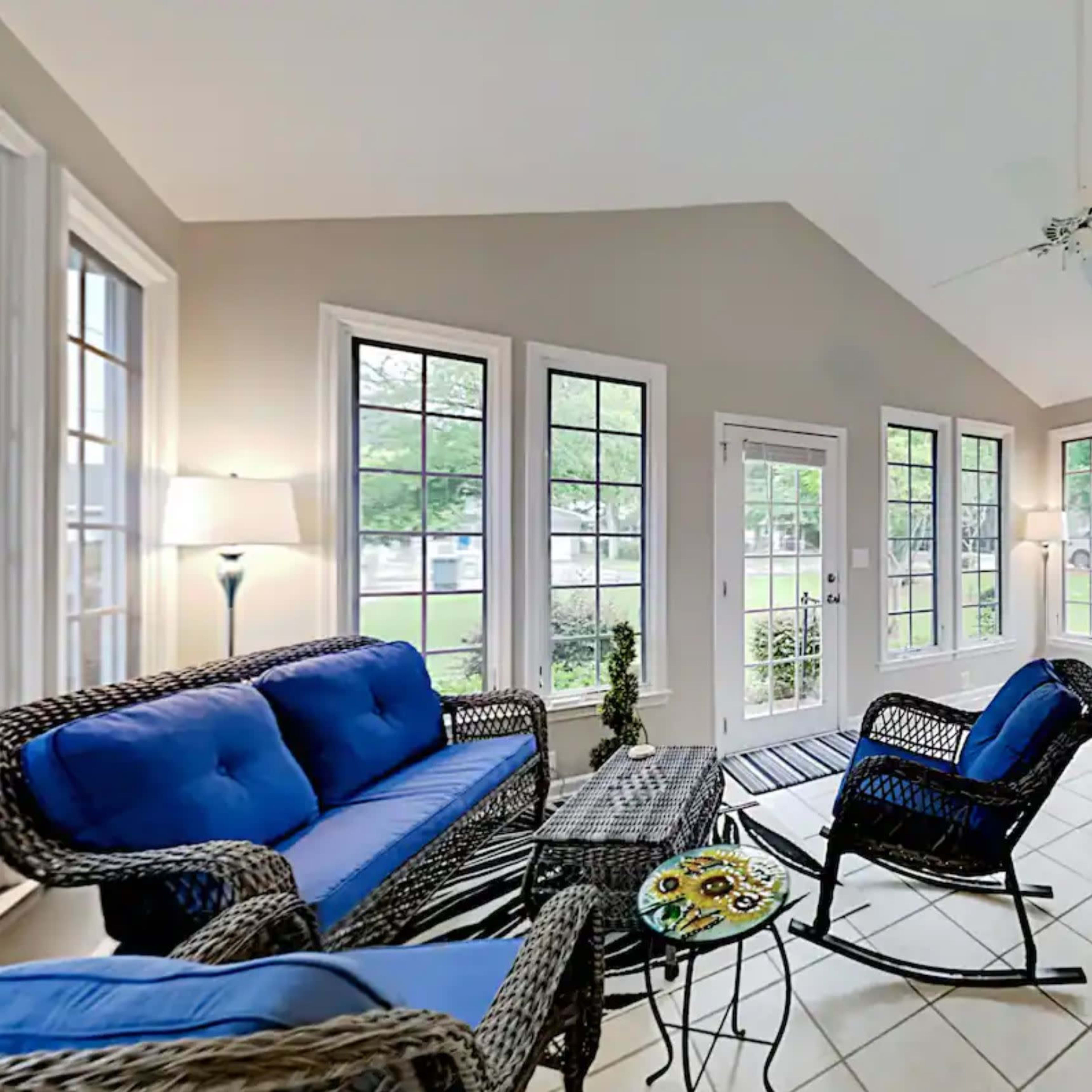 A sunlit sunroom featuring blue-cushioned wicker furniture, a dining table, and large windows overlooking a garden.