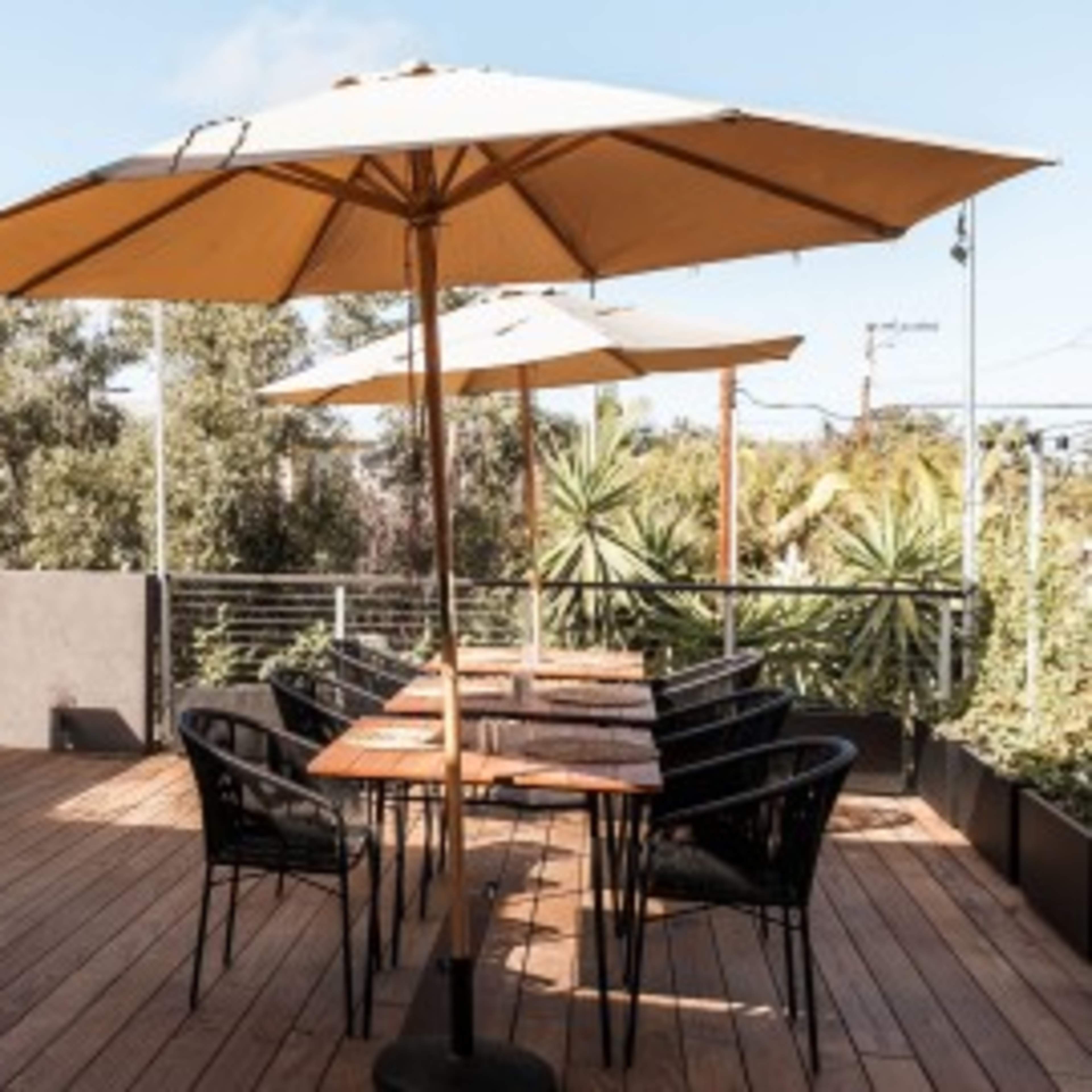 A rooftop terrace features wooden decking with a bar, high stools, dining tables, and large umbrellas surrounded by greenery.